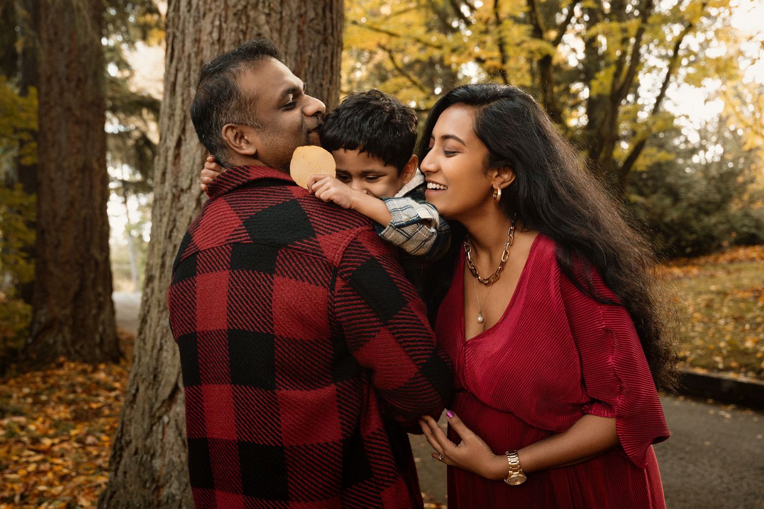 Family of three in red outfits shares a joyful moment outdoors among autumn foliage, child kissing father's cheek.