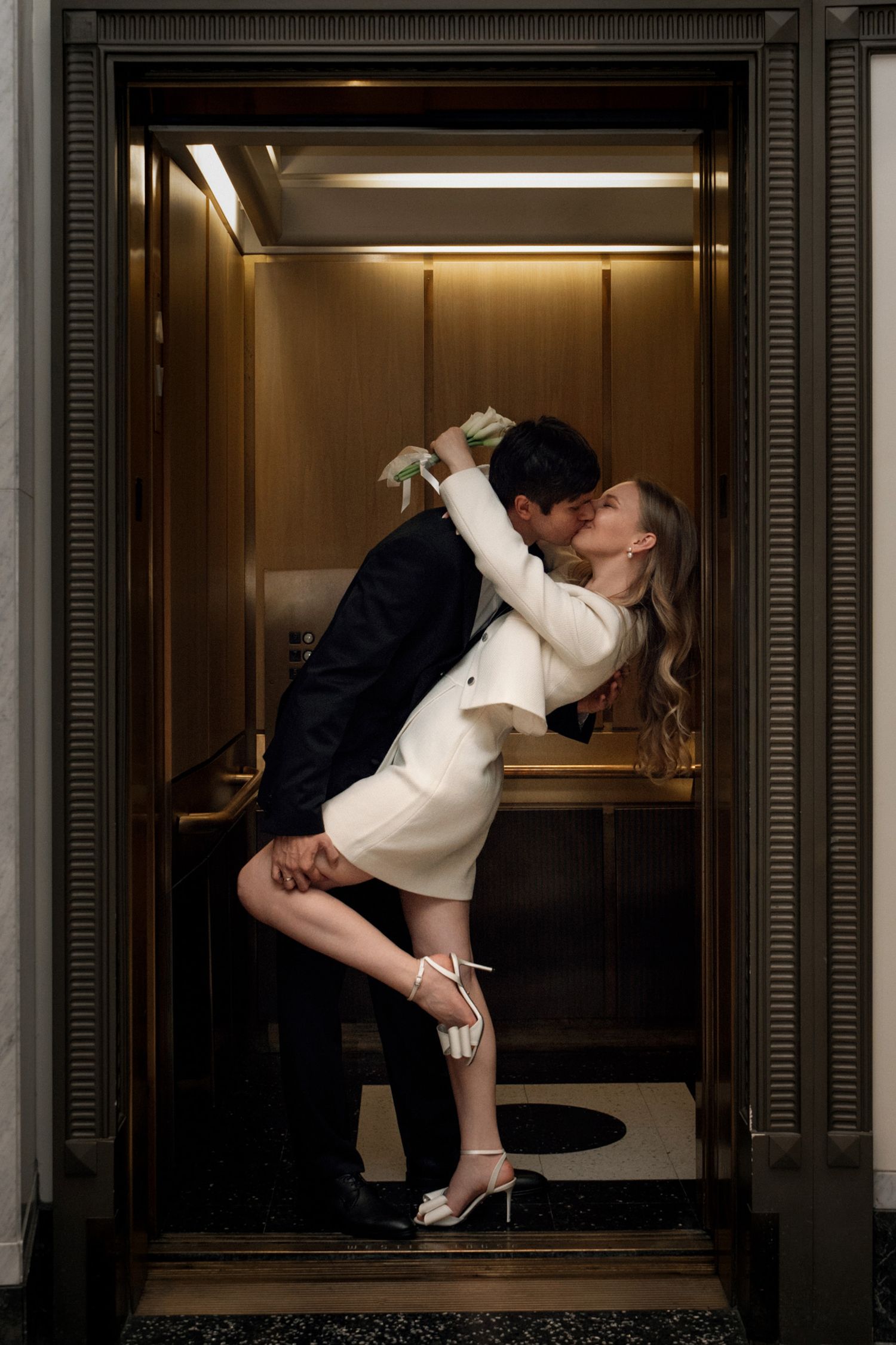 A couple shares a romantic dip kiss inside a golden elevator, bride in white mini dress and groom in dark suit.