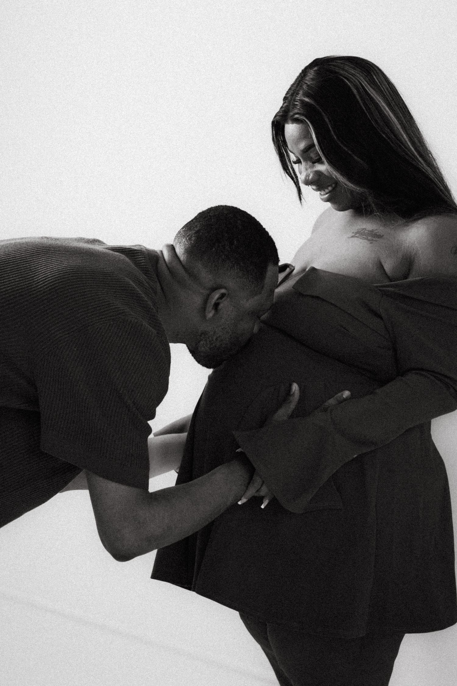 Black and white maternity photo of a man kissing a pregnant woman's belly while she smiles warmly.