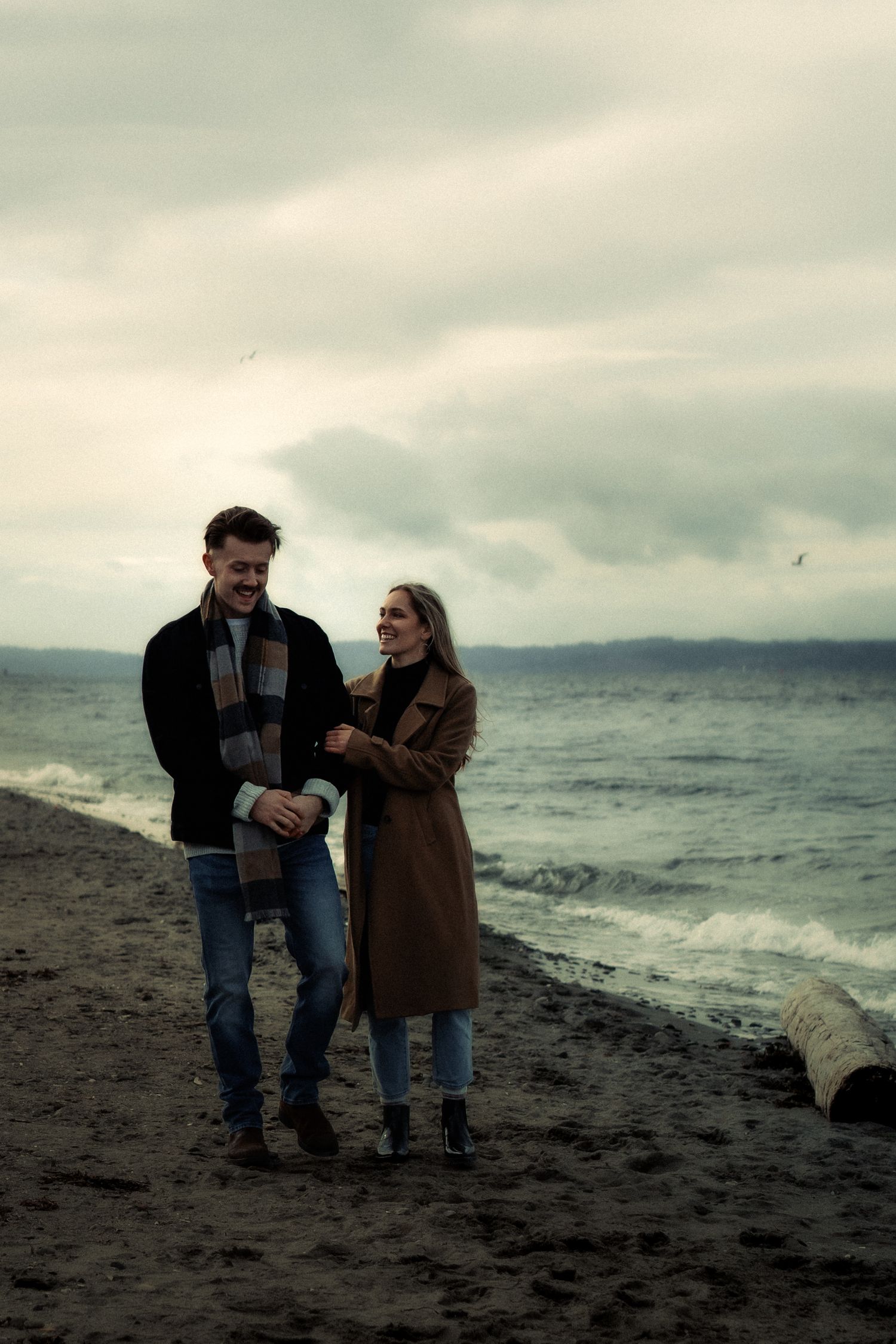 Couple walking hand in hand on a rocky beach on a cloudy day, wearing autumn coats and smiling at each other.