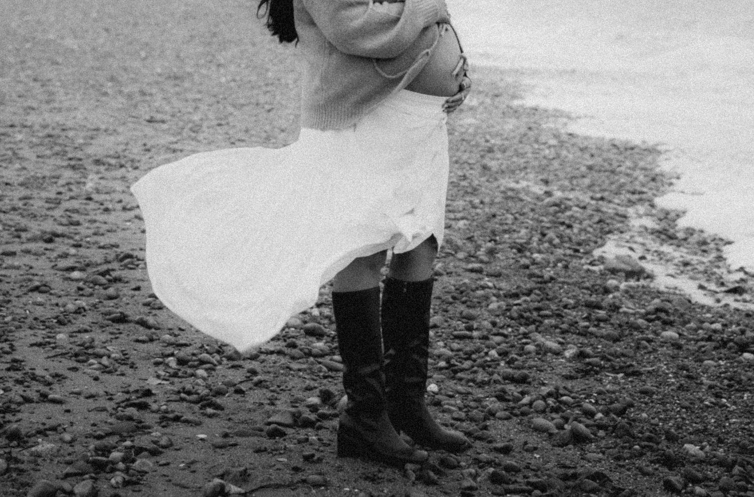 Black and white photo of a woman in a white dress and tall boots standing on a rocky beach in the wind.