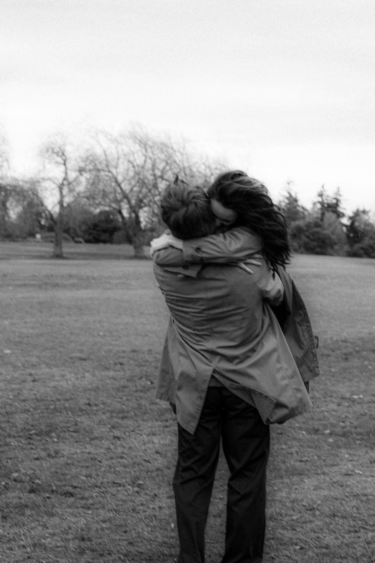 Black and white photo of a couple embracing in a park with bare trees in the background.