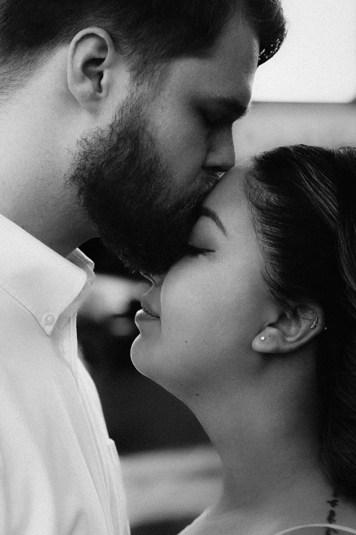 Black and white photo of a bearded man kissing a dark-haired woman on the forehead in a romantic moment.