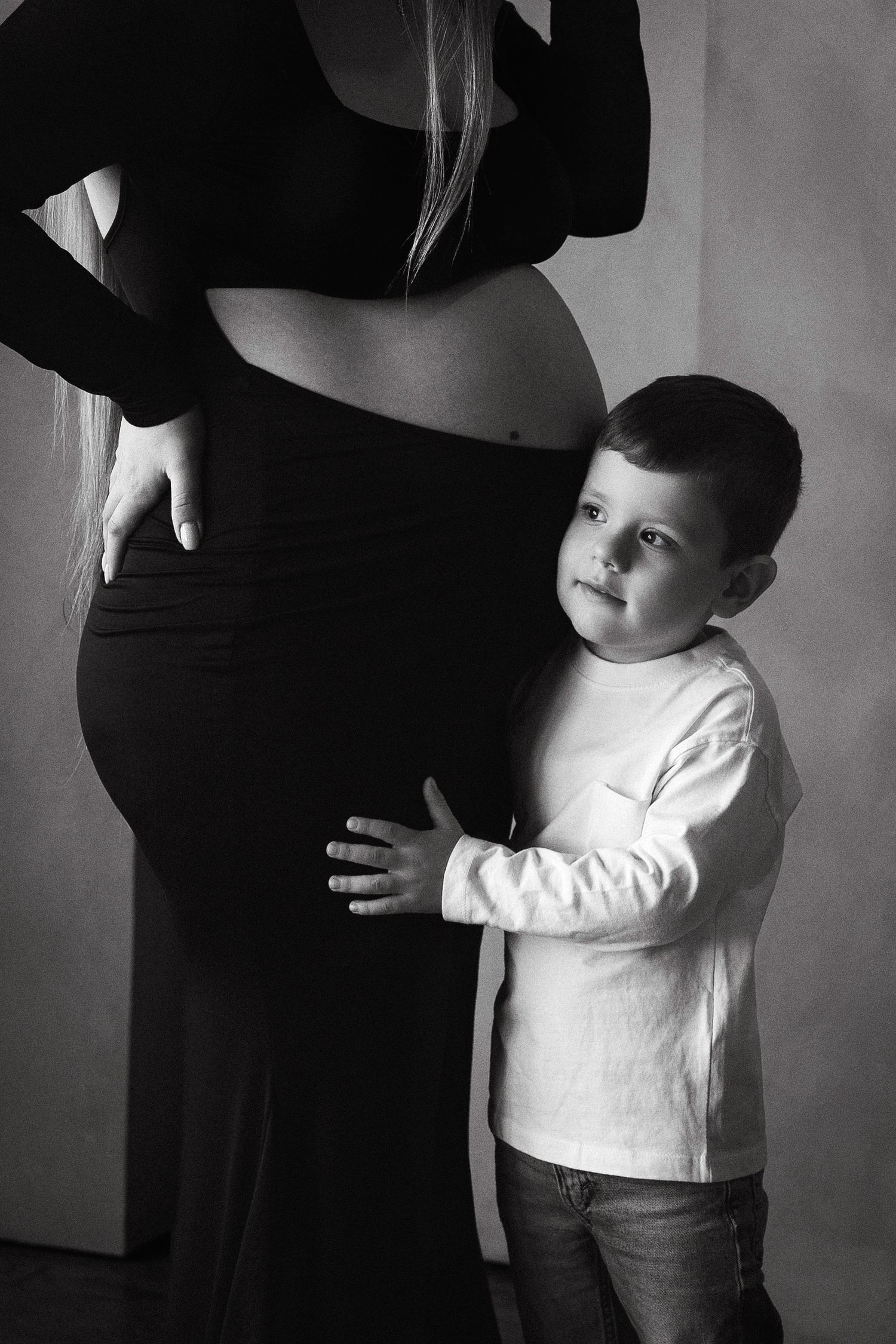 Black and white photo of a young boy hugging his pregnant mother's belly in a tender maternity portrait.