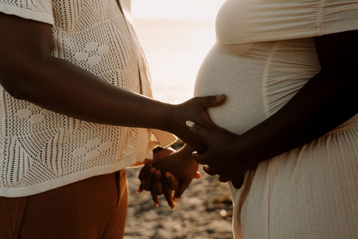 Two pregnant women holding hands at sunset on a beach, wearing white outfits, celebrating their pregnancies together.