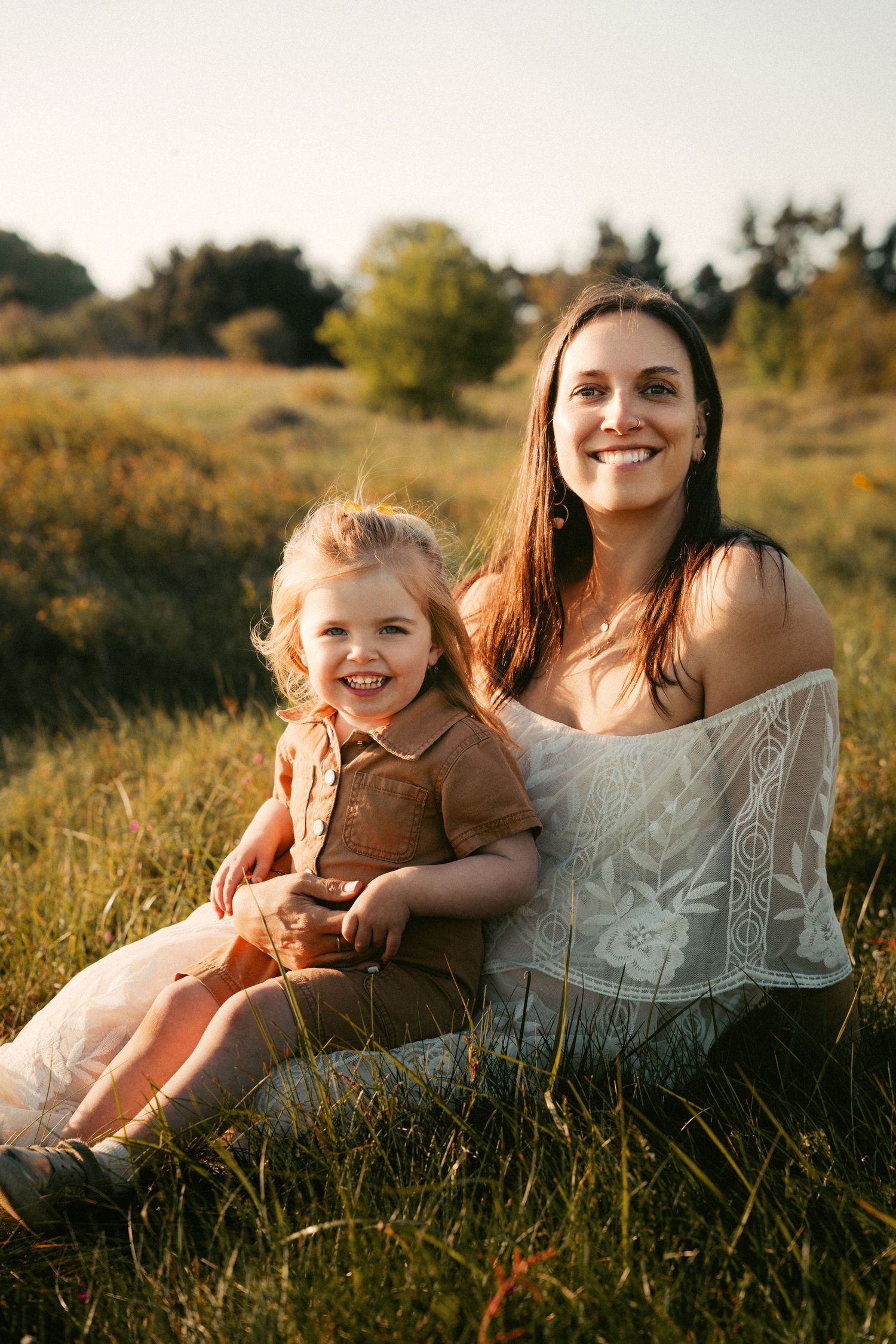 A smiling mother and young daughter sit together in a sunny golden field, wearing white and brown dresses.