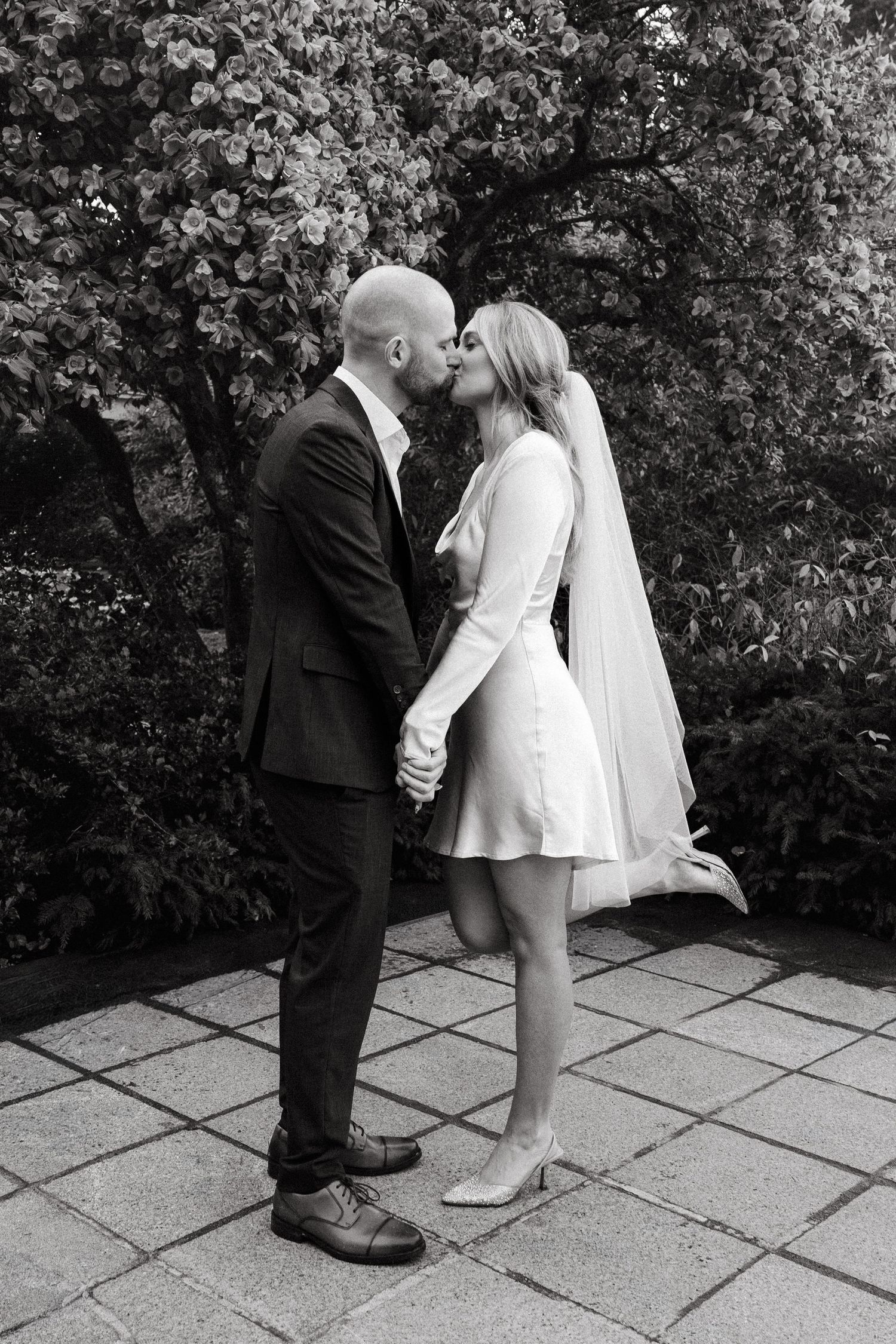 Black and white photo of a bride and groom sharing a kiss in a garden, holding hands in wedding attire.