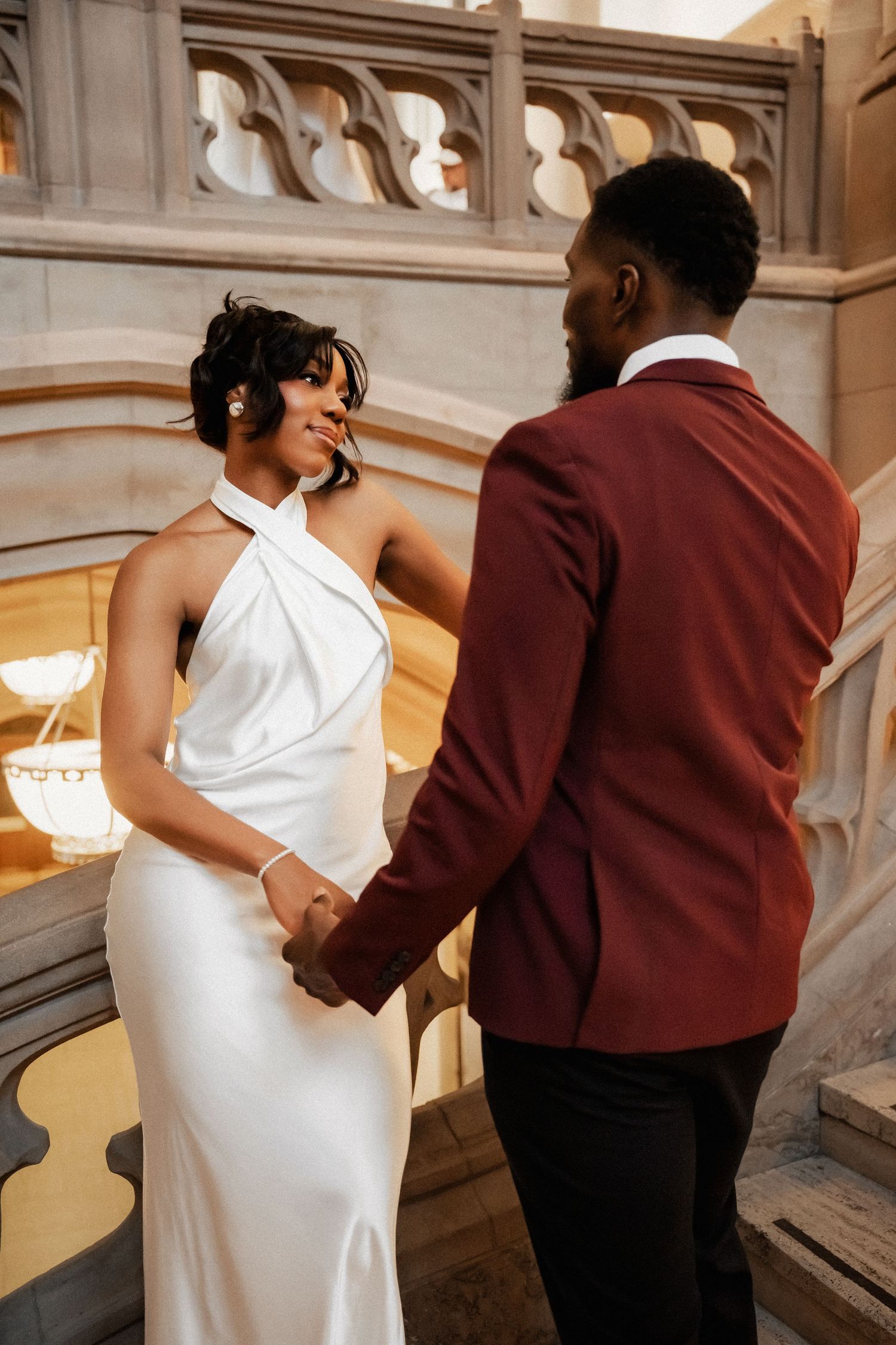 A couple in wedding attire holds hands on elegant stone stairs, the bride in white halter gown and groom in burgundy suit.