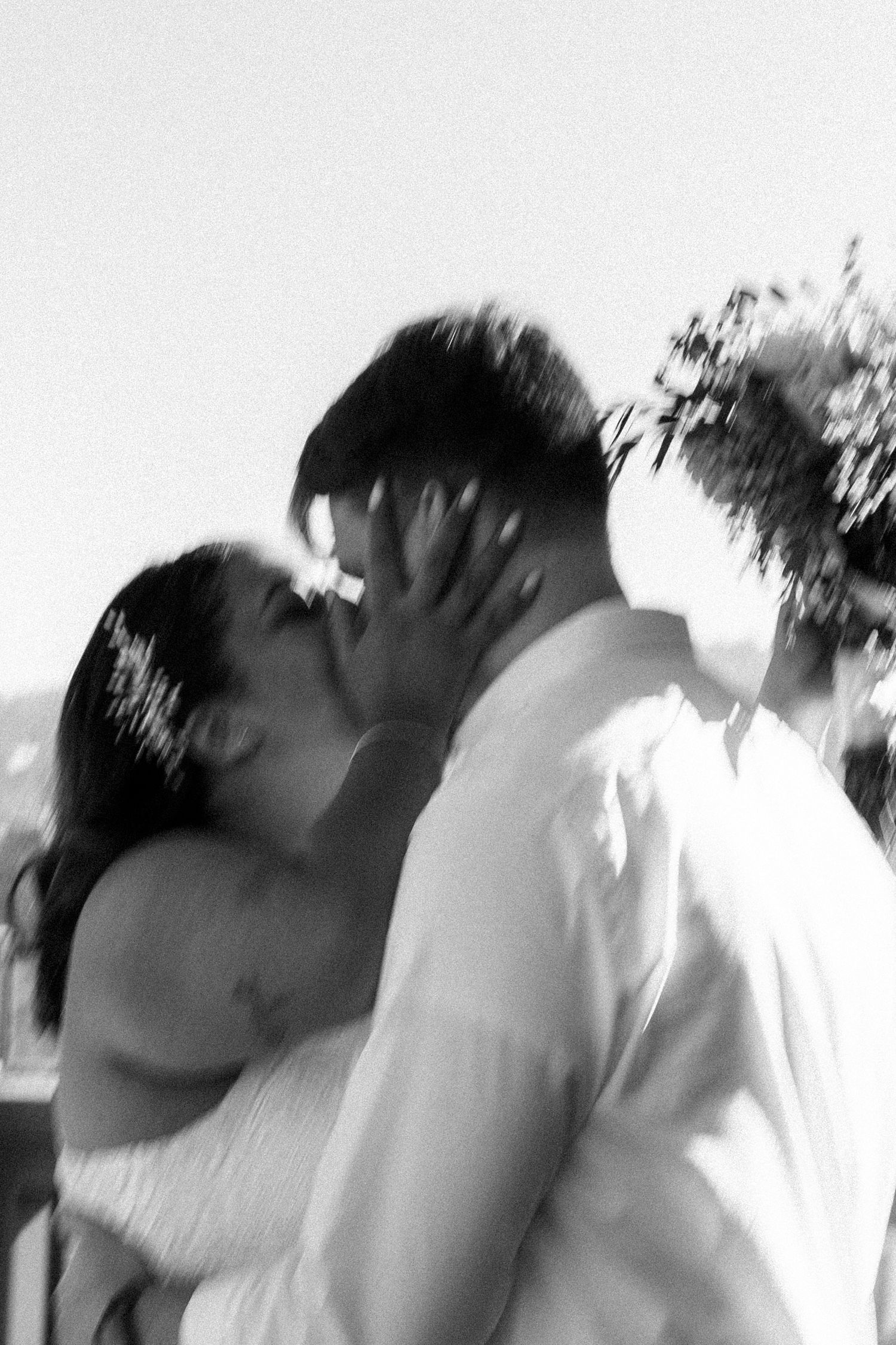 Black and white photo of a couple sharing a kiss outdoors, captured in a dreamy blurred motion effect.