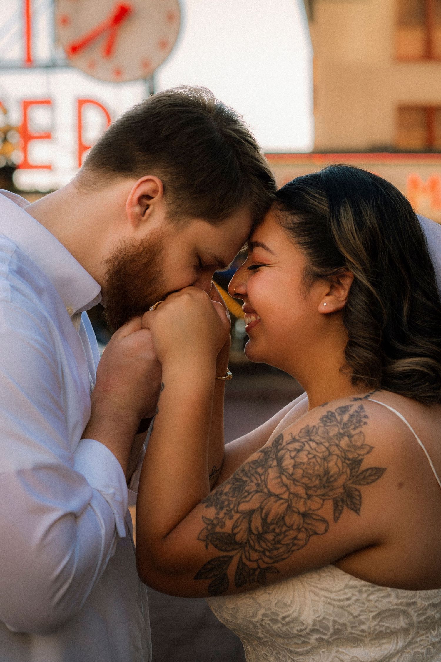 Couple sharing a romantic moment, forehead to forehead, woman with floral shoulder tattoo in lace dress, man in white shirt.