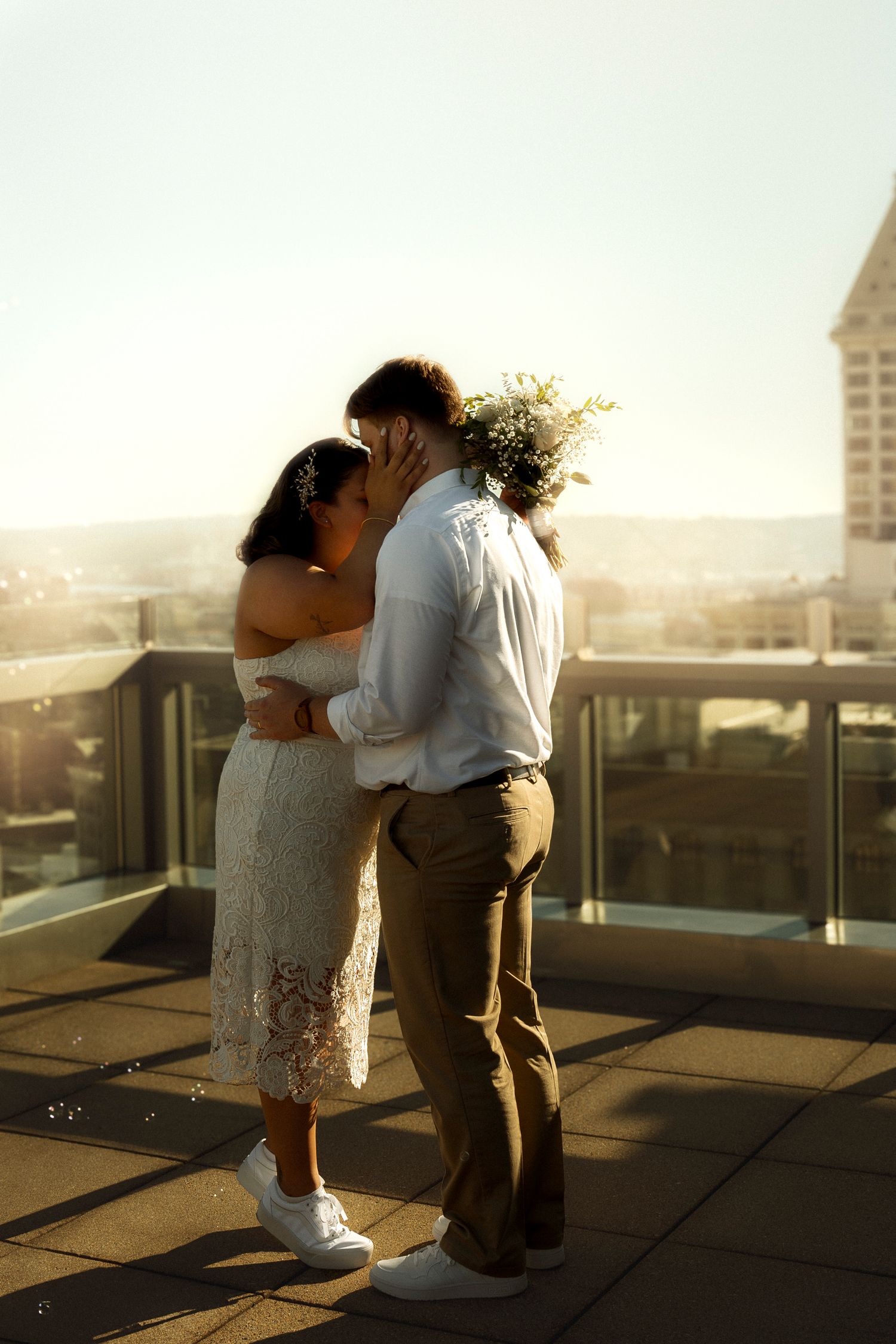 Couple sharing a romantic kiss on a rooftop terrace at golden hour, bride in white lace dress holding a bouquet.