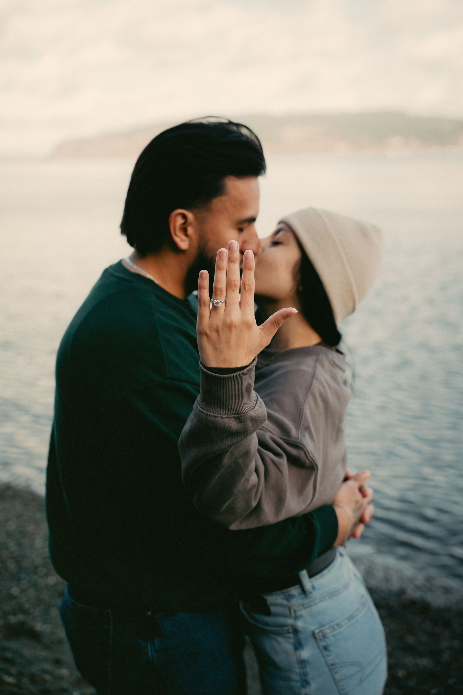 Couple sharing a romantic kiss by the water, woman showing off engagement ring while wearing a beige hat.