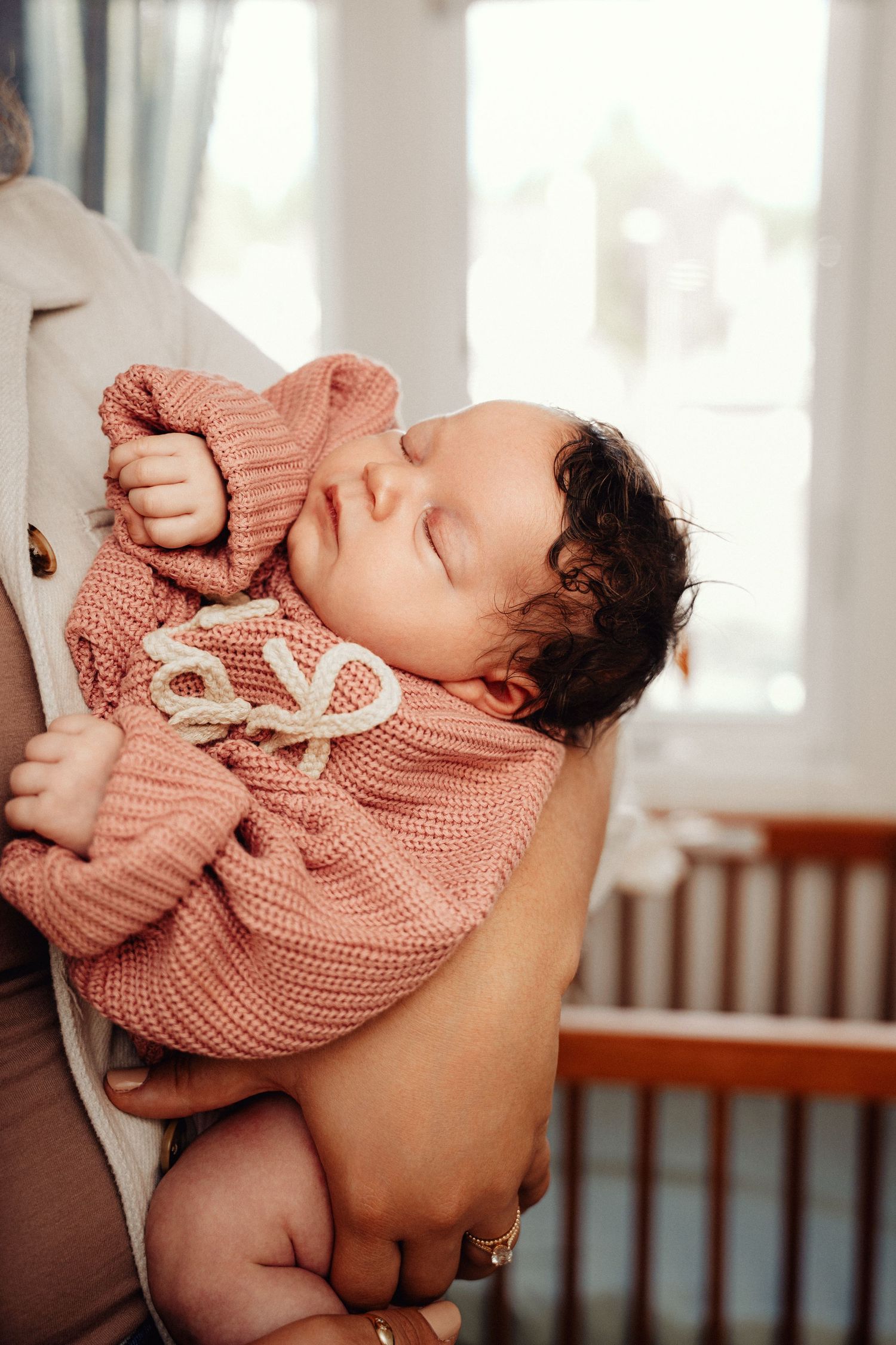 Newborn baby sleeping peacefully in parent's arms, wearing a pink knit sweater with bow detail.
