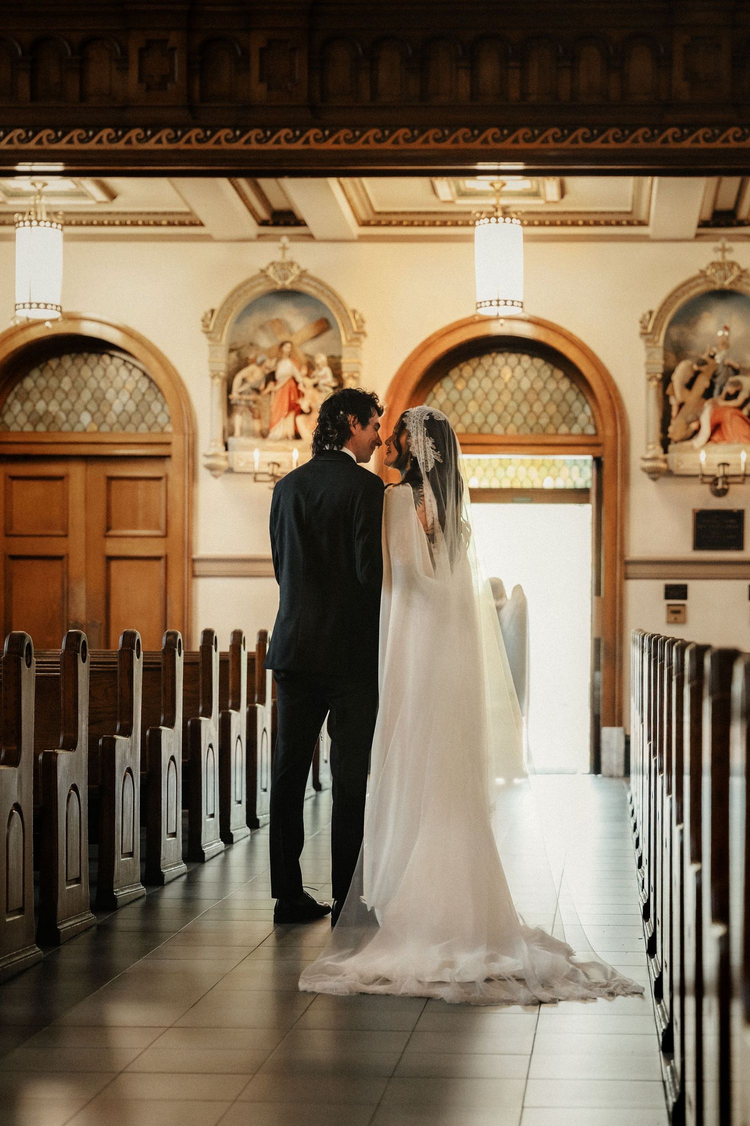 A bride and groom share a kiss at the altar of an elegant church with wooden pews and ornate arched windows.