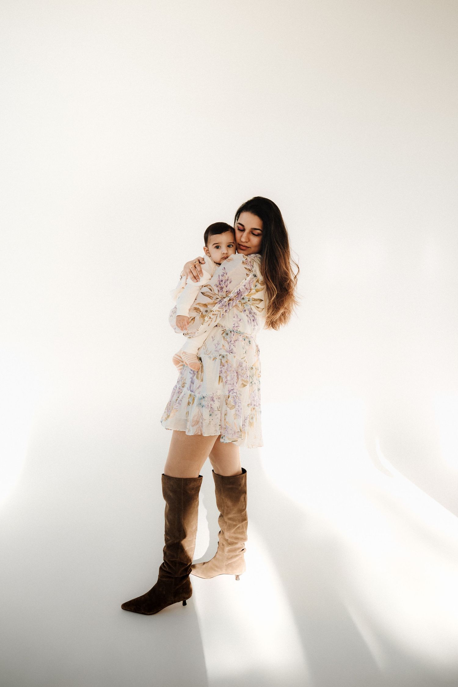 A stylish mother in a floral mini dress and brown knee-high boots holds her baby in a bright white studio.