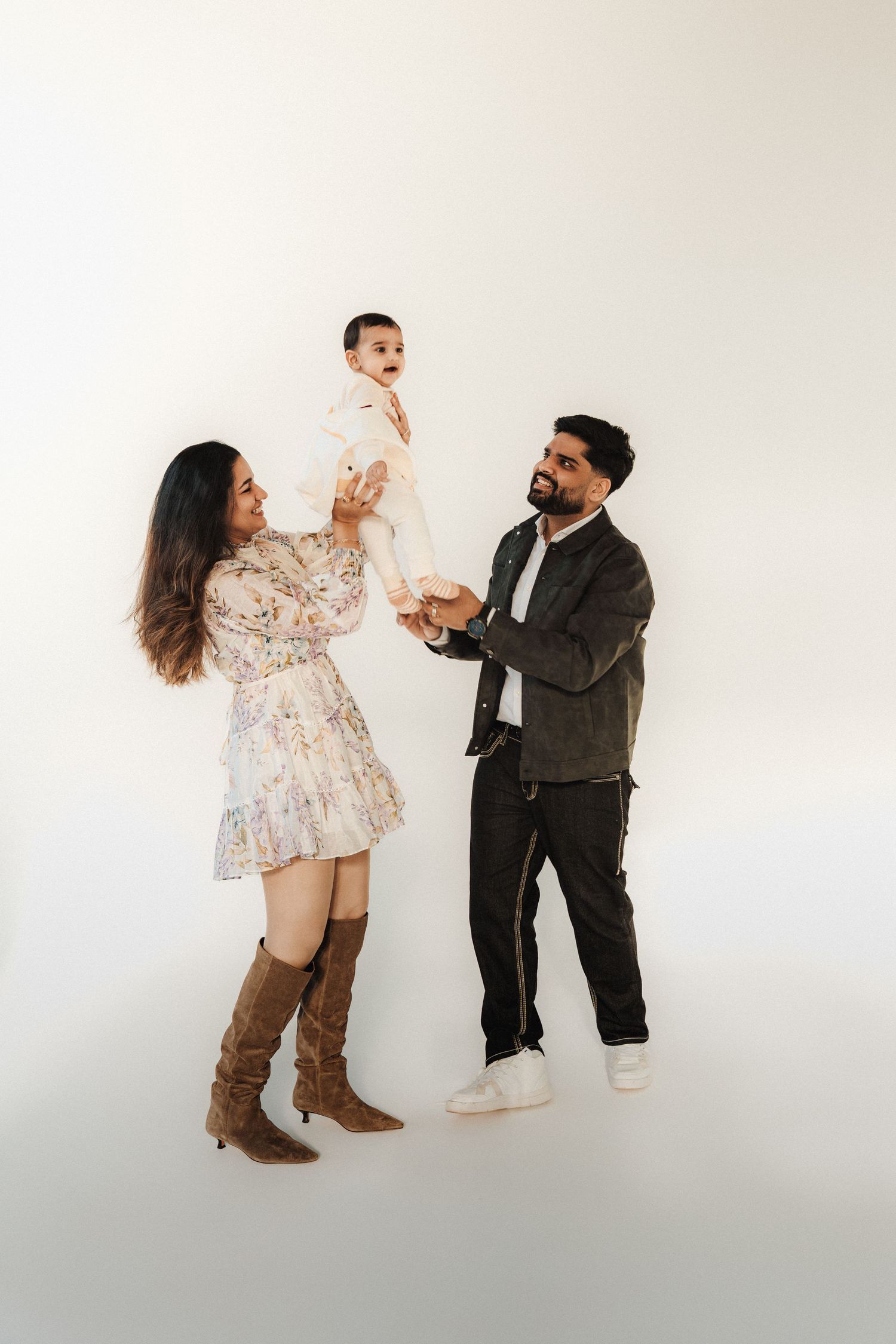 Happy family portrait with parents lifting smiling baby in white outfit against light background.