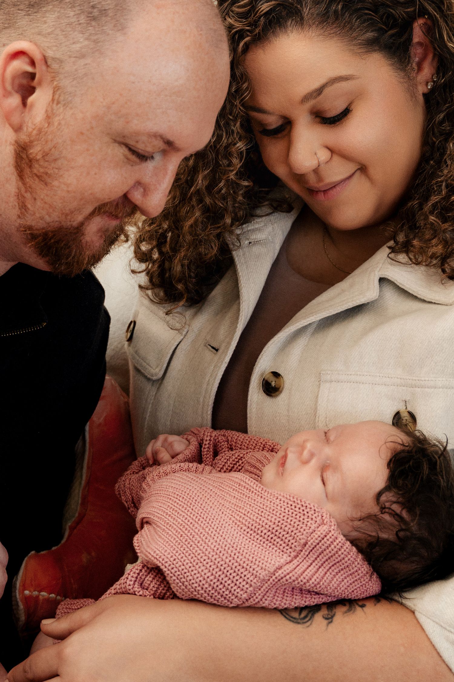 A couple smiles lovingly at their newborn baby wrapped in a pink knit blanket.