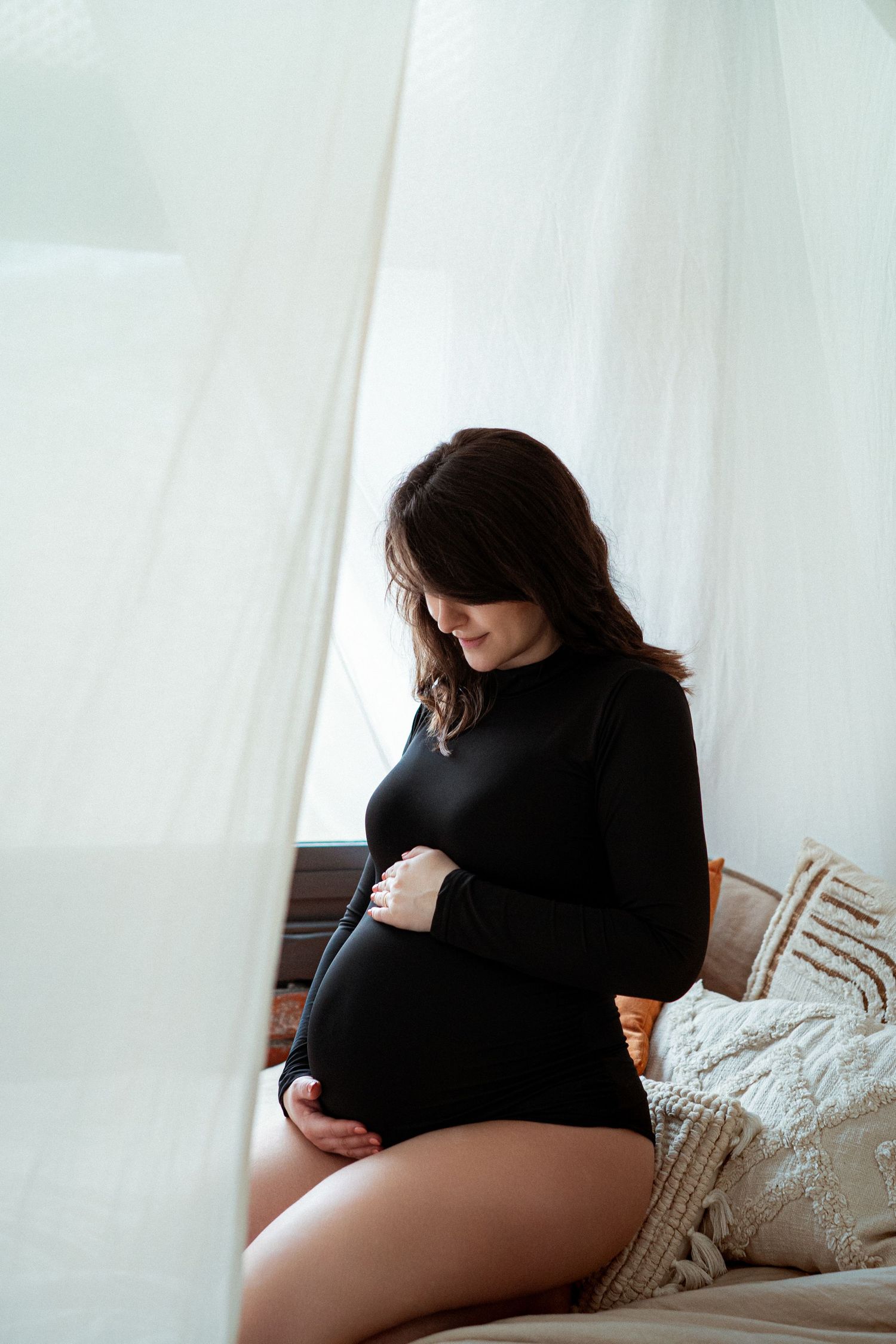 Pregnant woman in black dress cradling her baby bump while sitting on a bed near sheer white curtains.