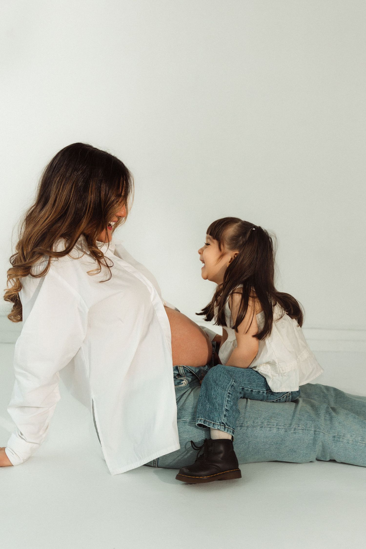 Pregnant woman in white shirt sitting on floor with young daughter in jeans, sharing a joyful moment together.