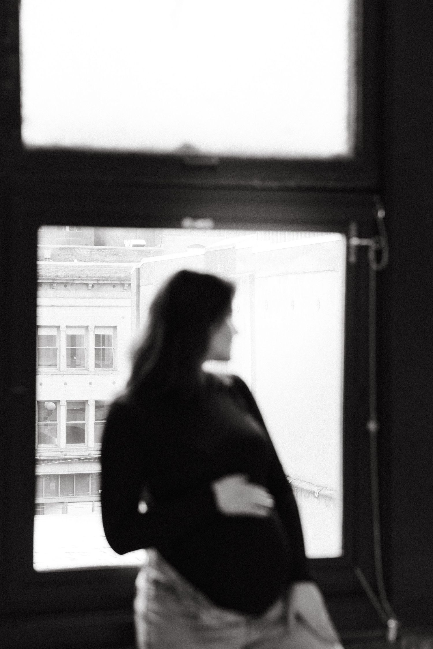 Black-and-white photo of a pregnant woman standing by an urban window, cradling her baby bump in a soft silhouette. Seattle and Tacoma