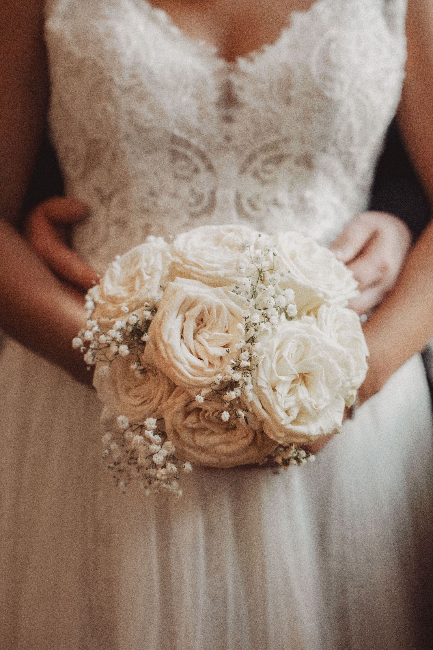 Bride holding a round bridal bouquet of white roses and baby's breath, wearing a lace wedding dress.