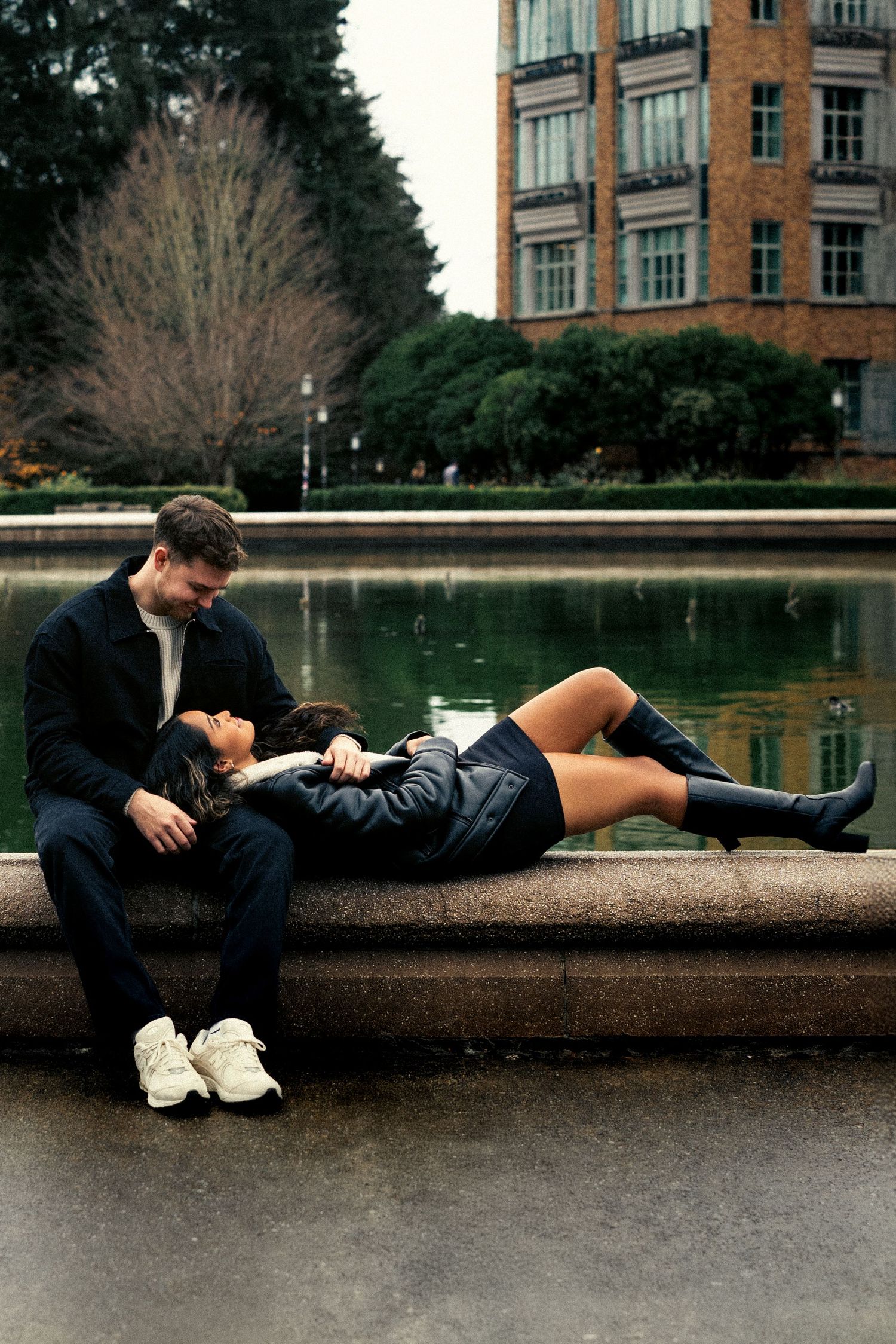 Couple sitting by a fountain in a park, woman reclining on man's lap, both dressed in black, urban background.