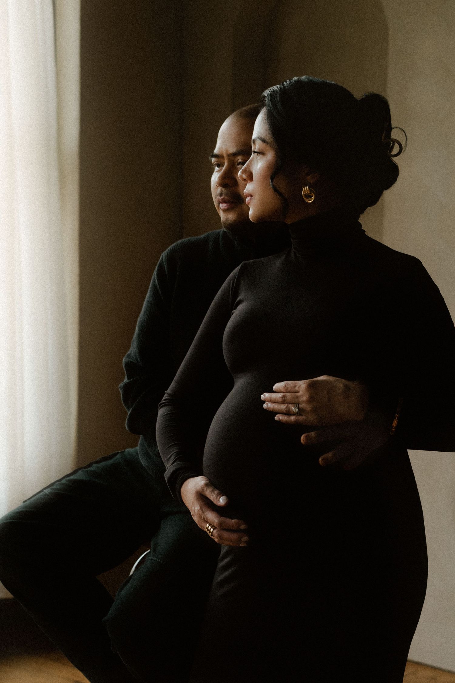 Couple in black outfits sharing a tender maternity moment, man embracing pregnant woman by a sunlit window.