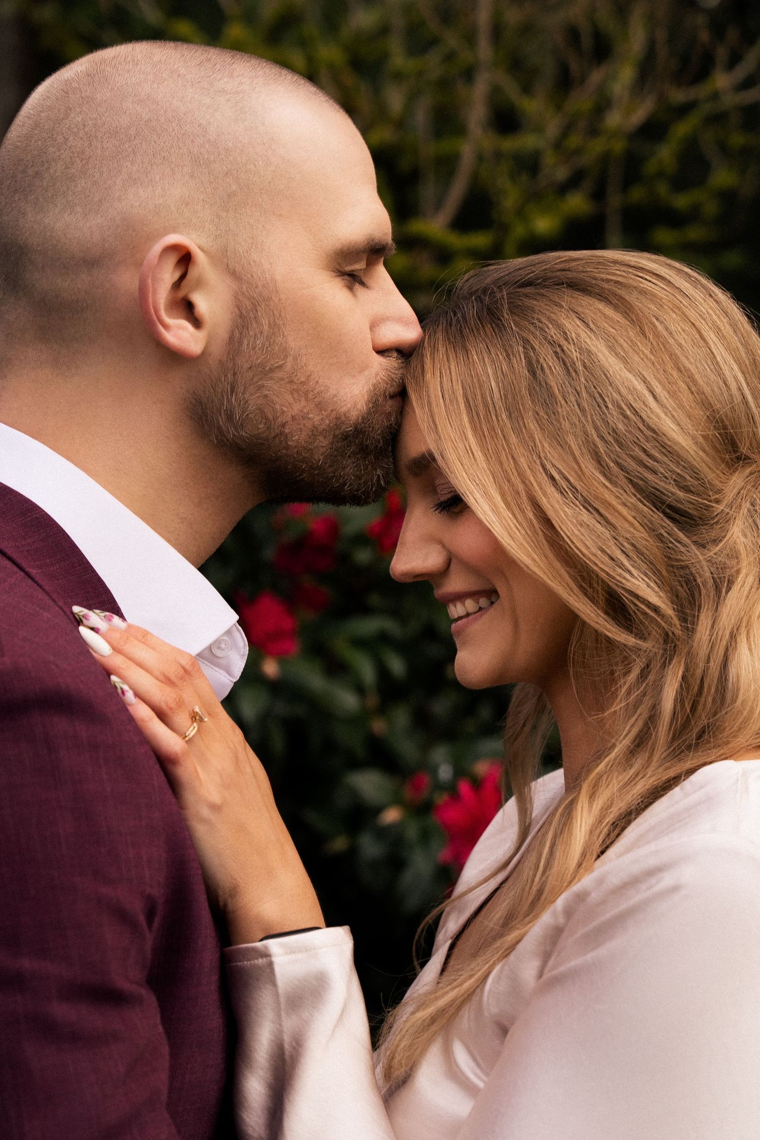 A bald man in a burgundy suit kisses a smiling blonde woman on the forehead in a romantic garden setting.