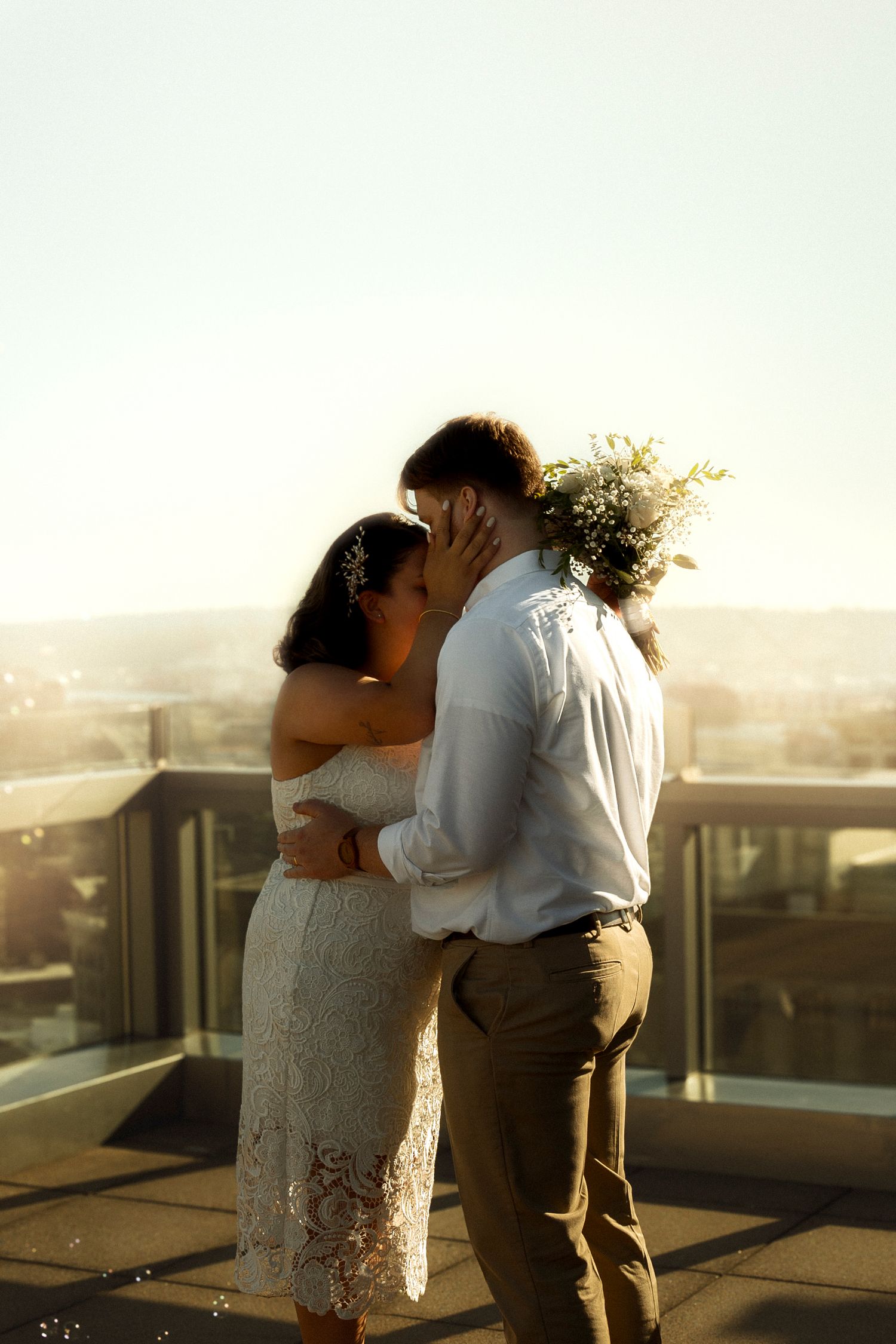 Bride and groom sharing a romantic kiss on a rooftop at golden hour, bride holding a lush bouquet.