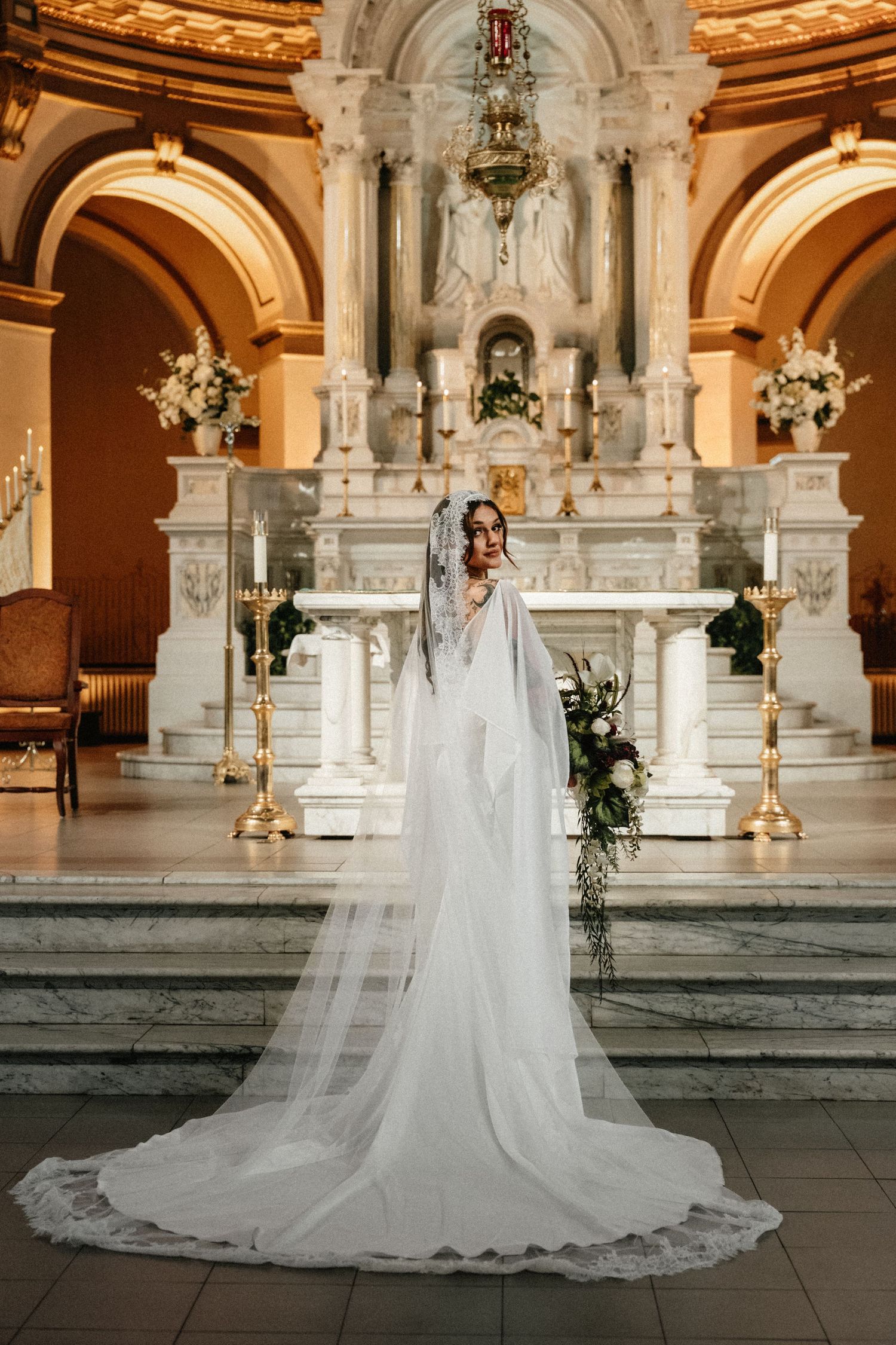 Bride in white gown and long veil stands before ornate marble altar in elegant church setting.