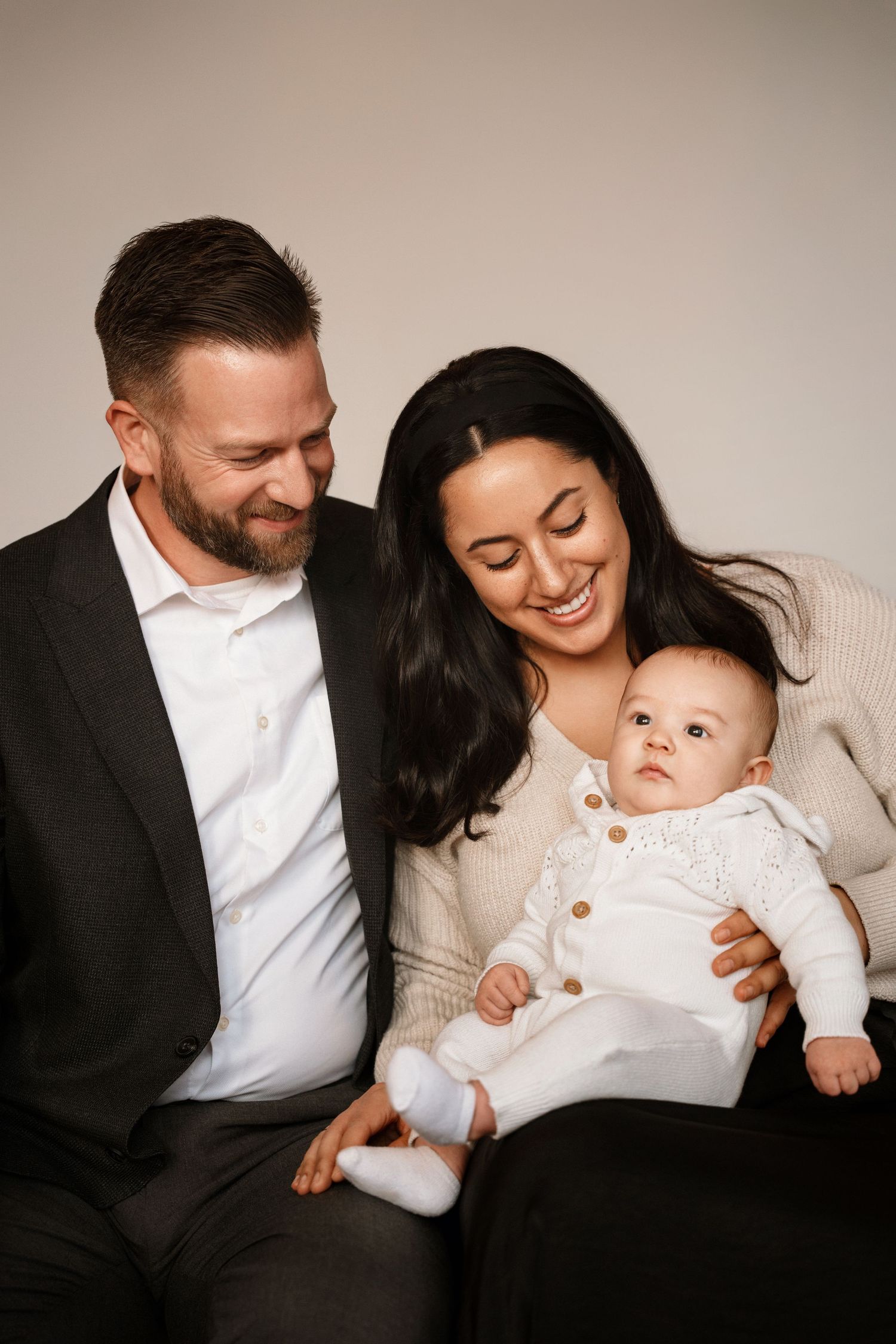 A smiling couple in a studio portrait gazes lovingly at their baby dressed in white, creating a warm family moment.
