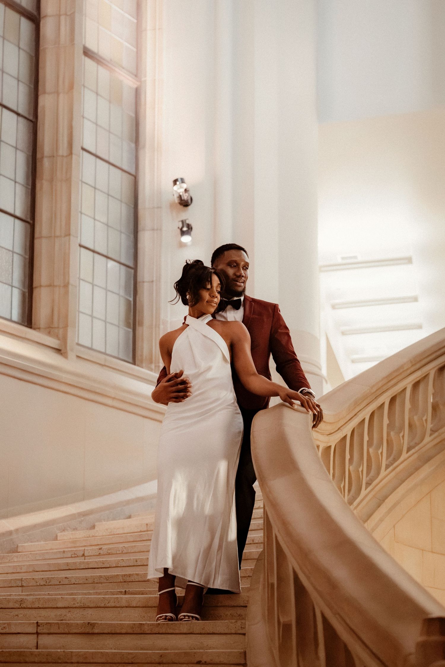 Elegant couple in wedding attire posing on grand marble staircase, groom in burgundy suit, bride in white halter dress.