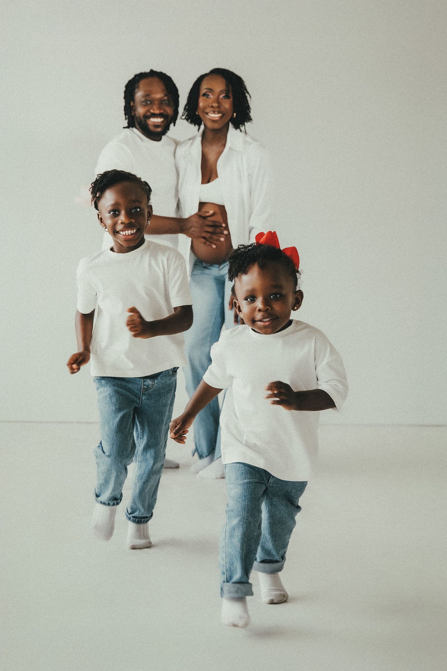 Happy Black family in matching white shirts and jeans, parents smiling as two young children run toward camera.