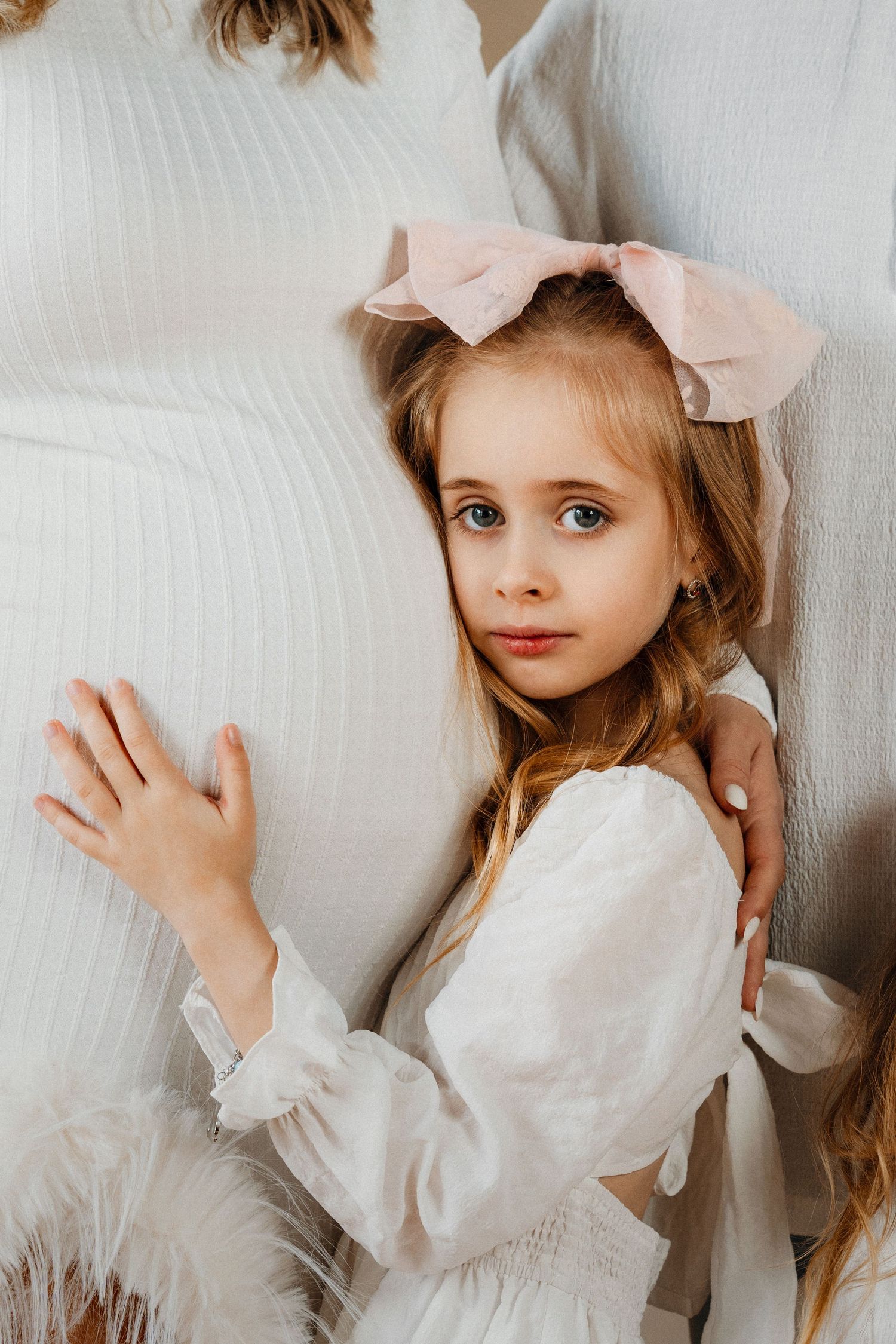 Young girl with long blonde hair and pink bow wearing white dress, leaning against cushions with a gentle gaze.