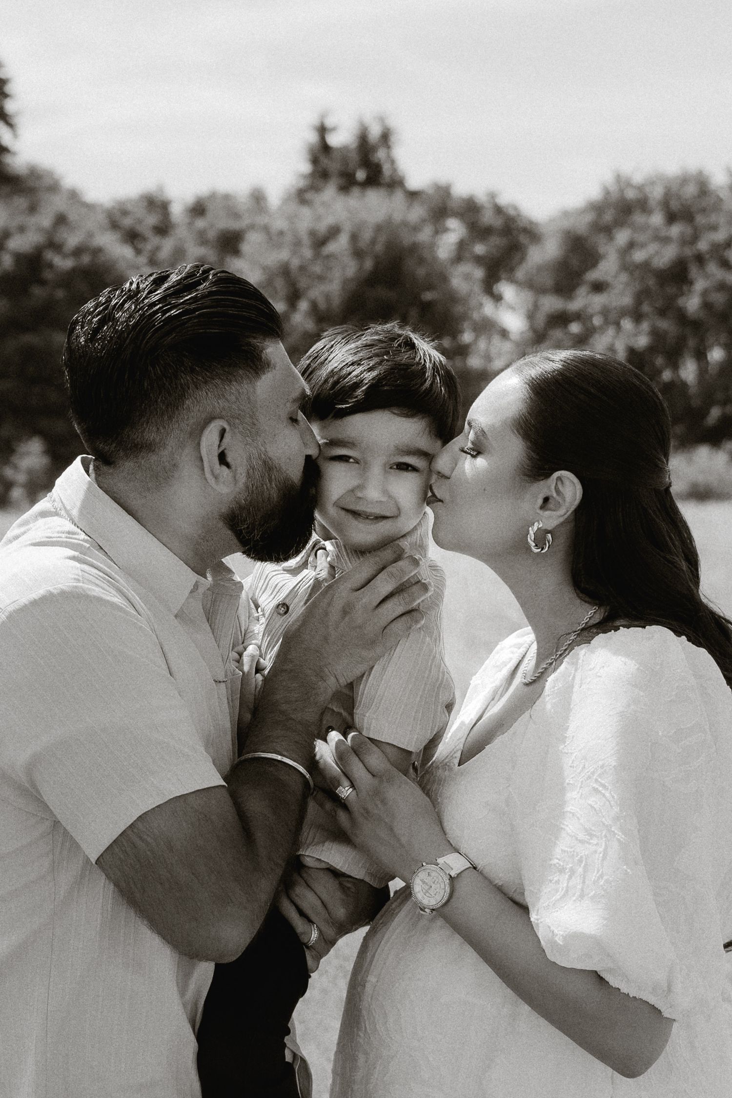 Black and white photo of parents kissing their smiling toddler on the cheeks outdoors.
