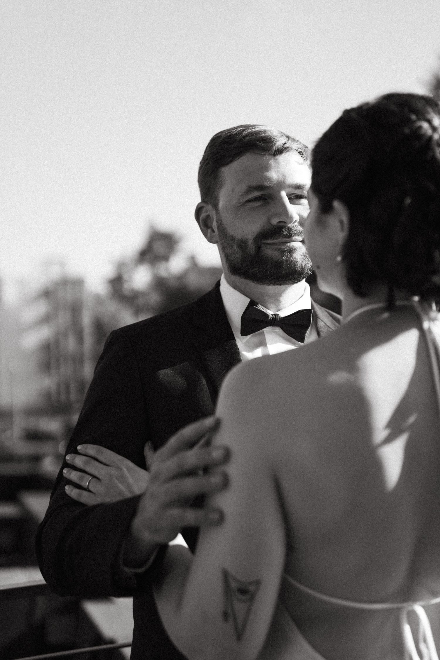 Smiling bearded groom in black tuxedo dancing with bride on sunny outdoor terrace.