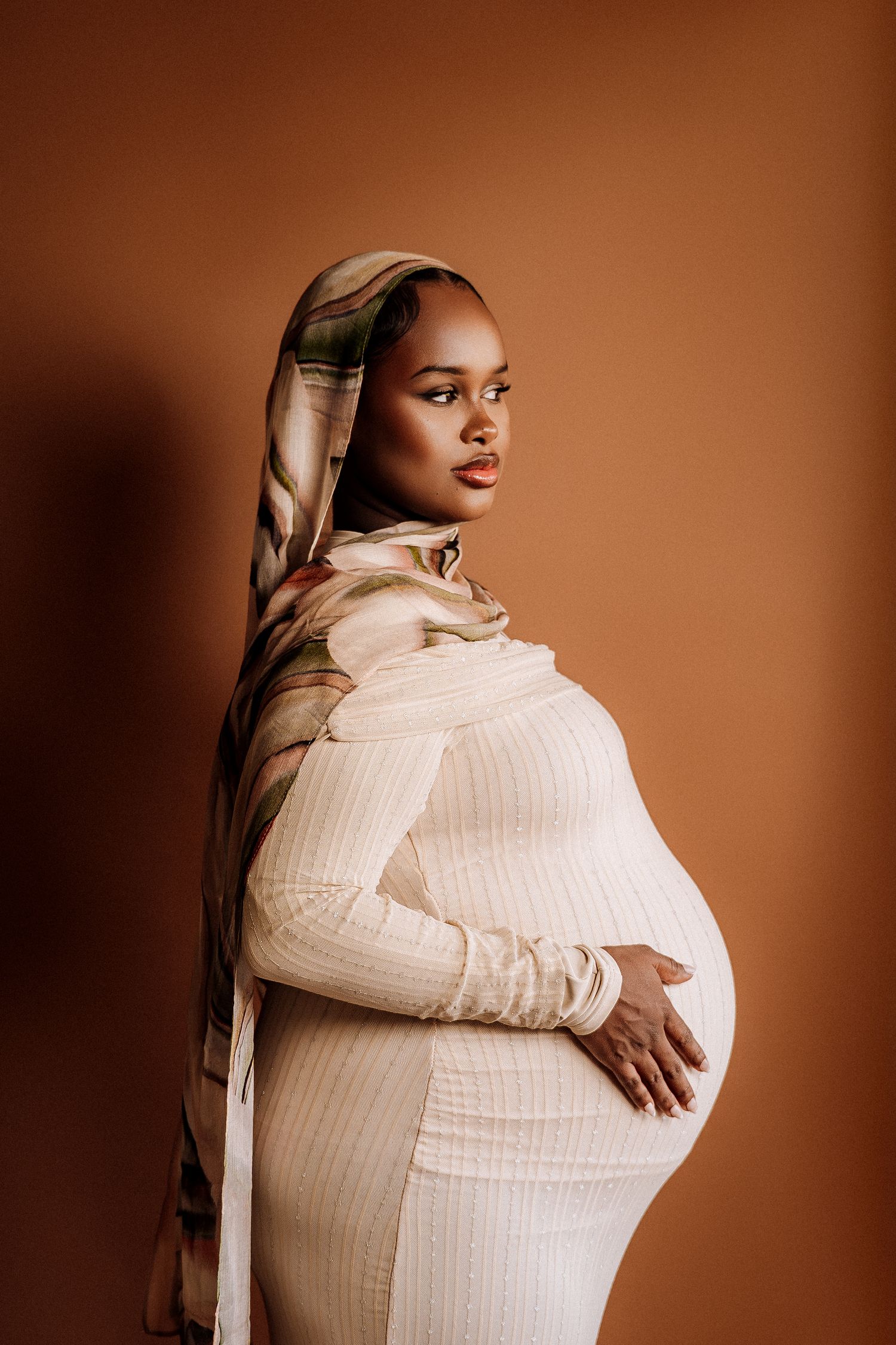 Pregnant woman in white ribbed dress and floral head wrap poses gracefully against warm brown background.