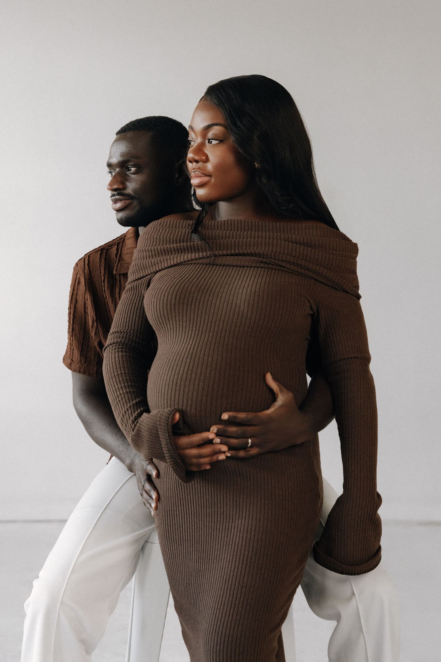 A pregnant Black woman in a brown ribbed dress is embraced from behind by a man in a brown shirt against a white background.