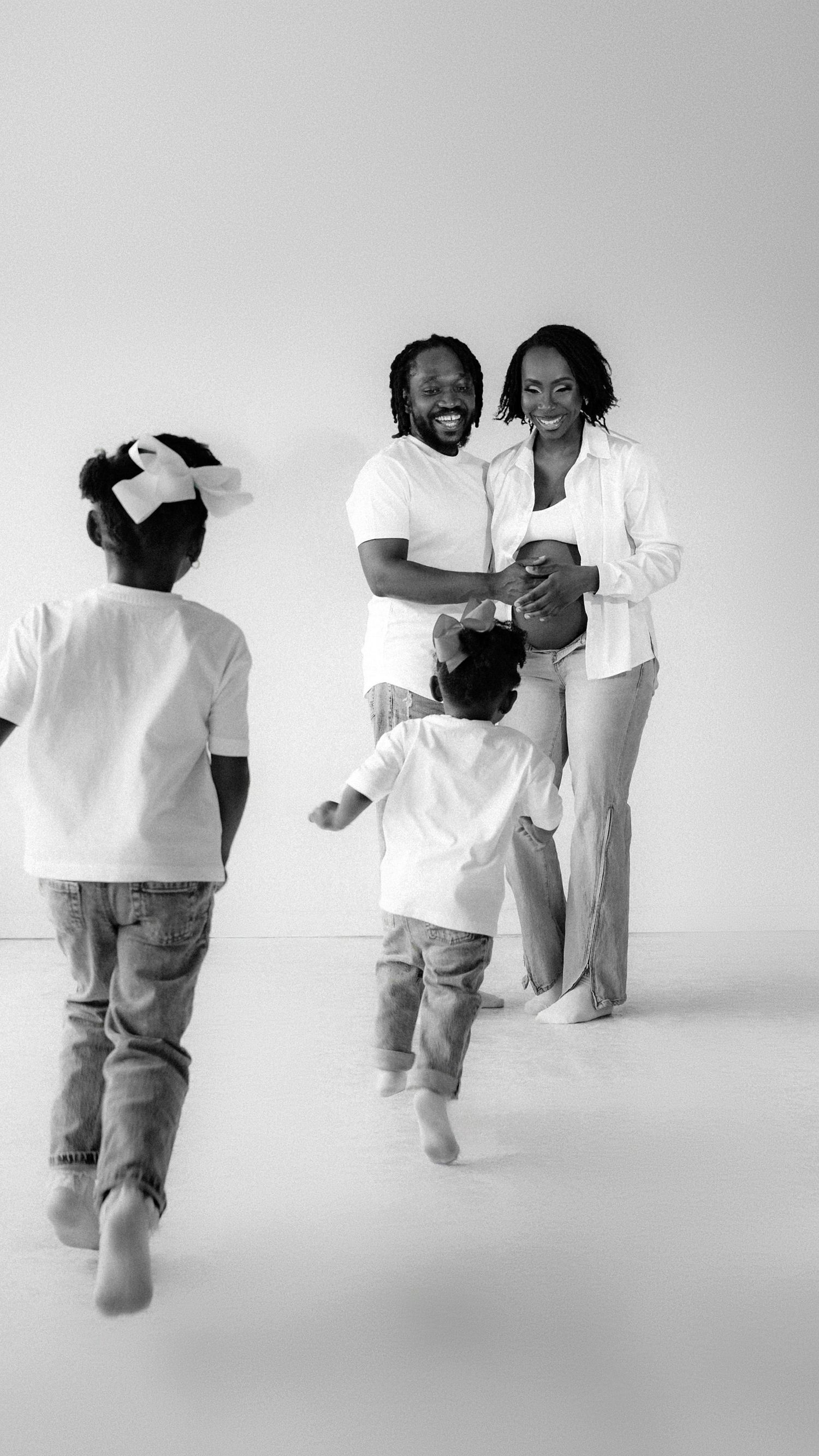 Black and white family photo of parents embracing while two young children run toward them in a bright white studio.