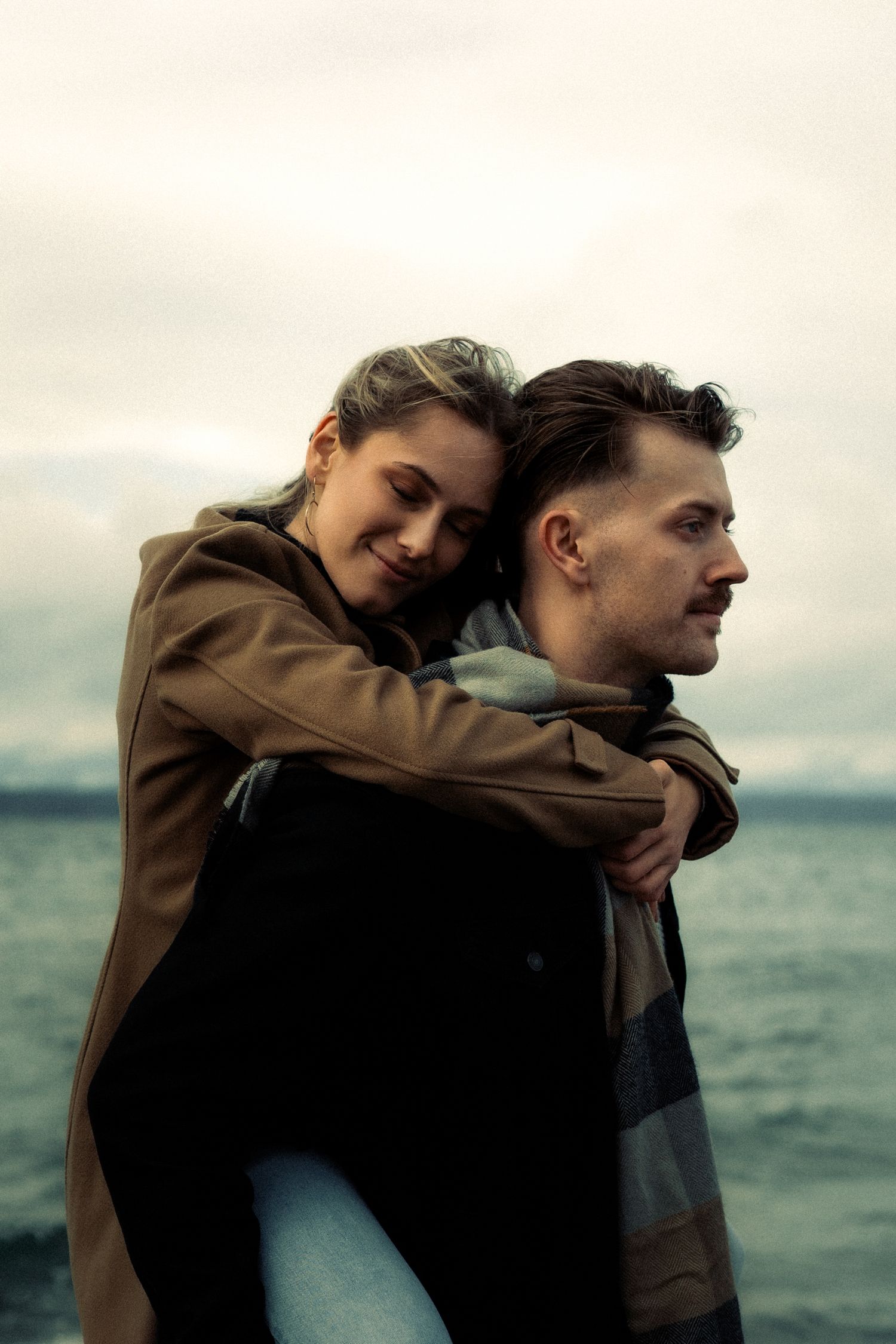 A couple shares a tender piggyback embrace by the ocean, woman smiling with eyes closed, both in casual autumn coats.