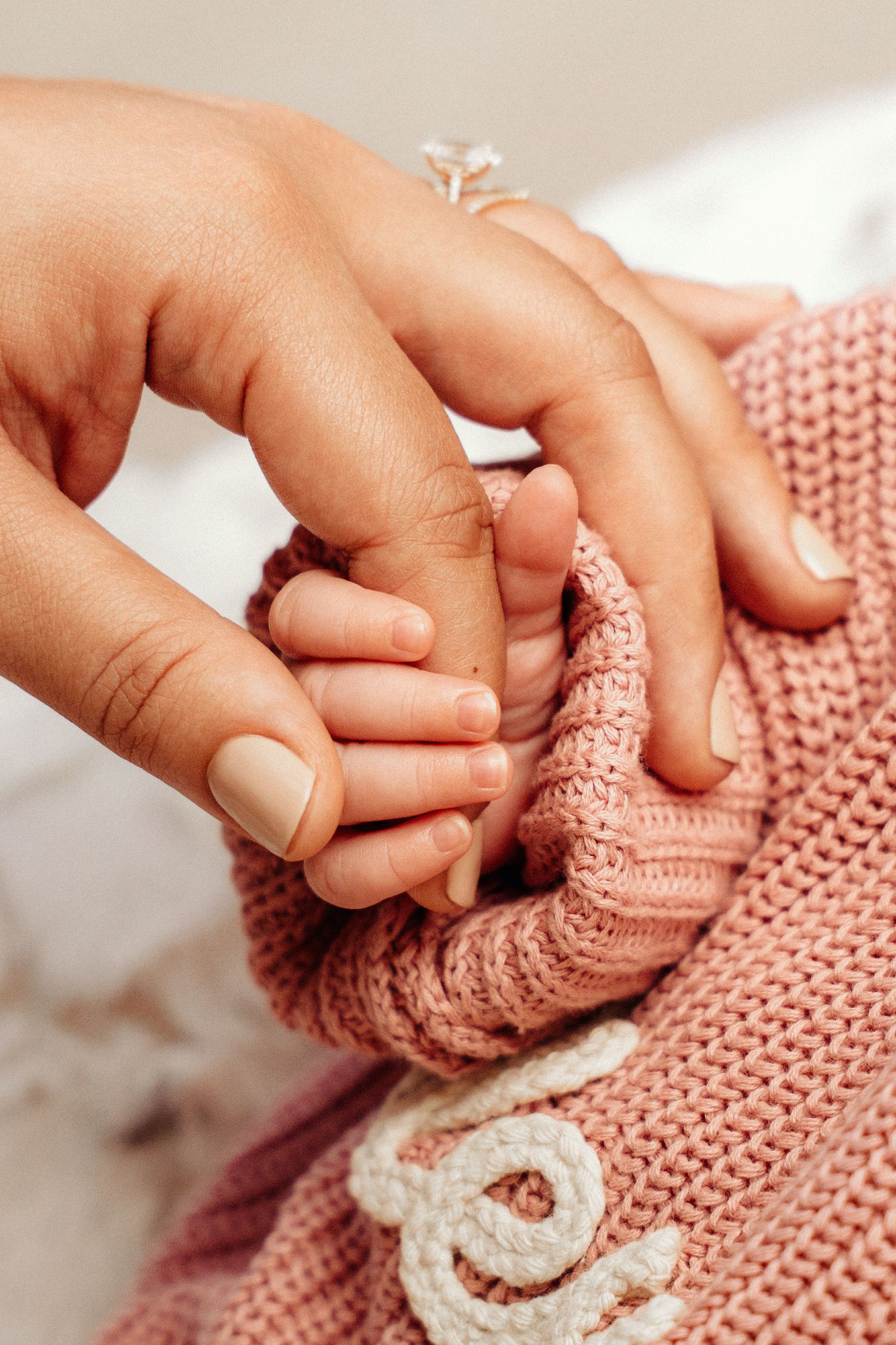 A mother gently holds her newborn baby's tiny hand, wrapped in a soft pink knitted blanket.