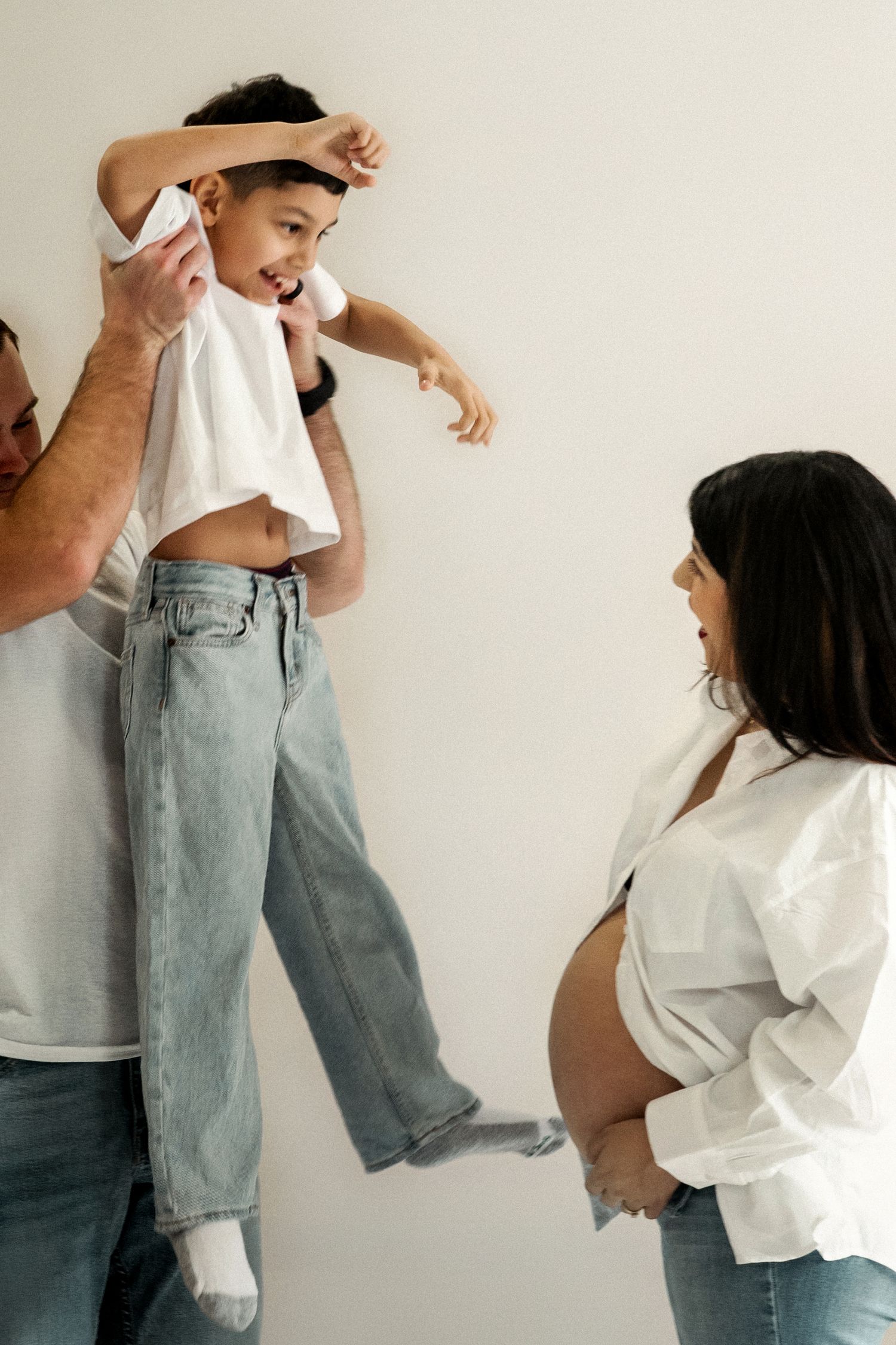 Pregnant woman smiling at child being lifted by partner during a fun family maternity photo session.