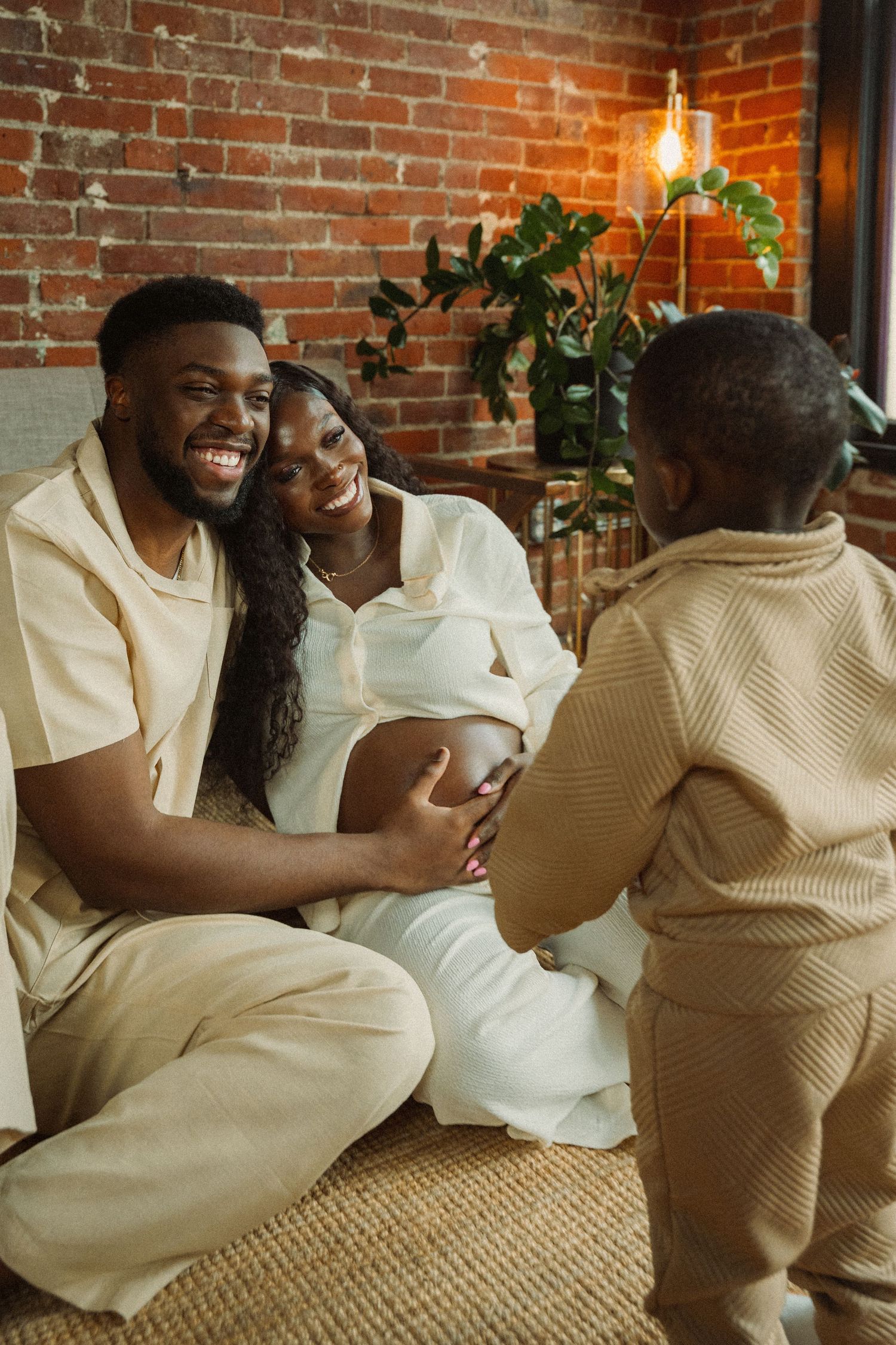 A joyful Black family with a pregnant mother, smiling father, and toddler sharing a warm moment in a cozy brick-walled room.