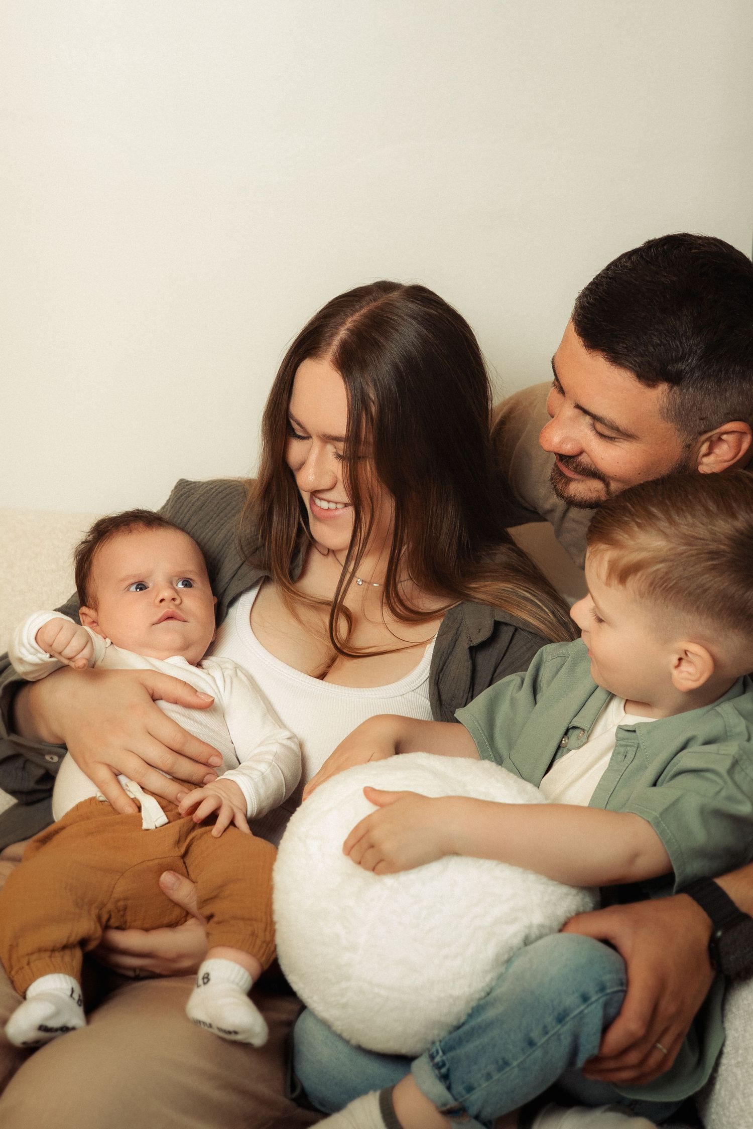 Happy family of four sitting together on a couch, parents smiling at their newborn baby and young toddler son.