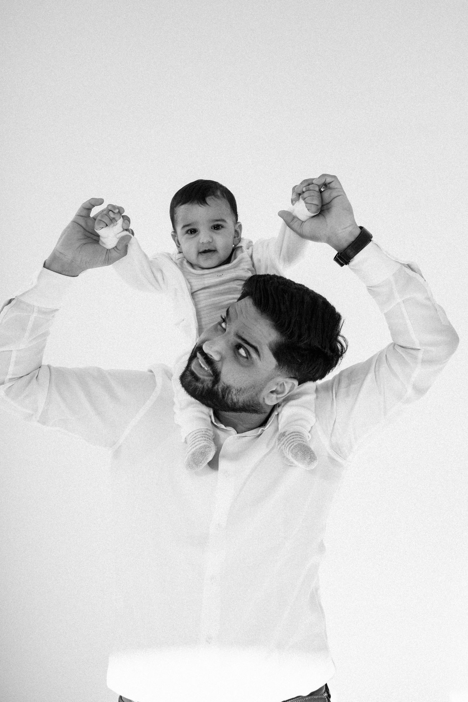 Black and white photo of a bearded father holding his baby on his shoulders against a white background.