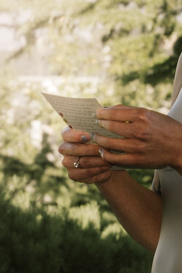 Close-up of hands holding handwritten wedding vows outdoors, with rings visible against a lush green background.