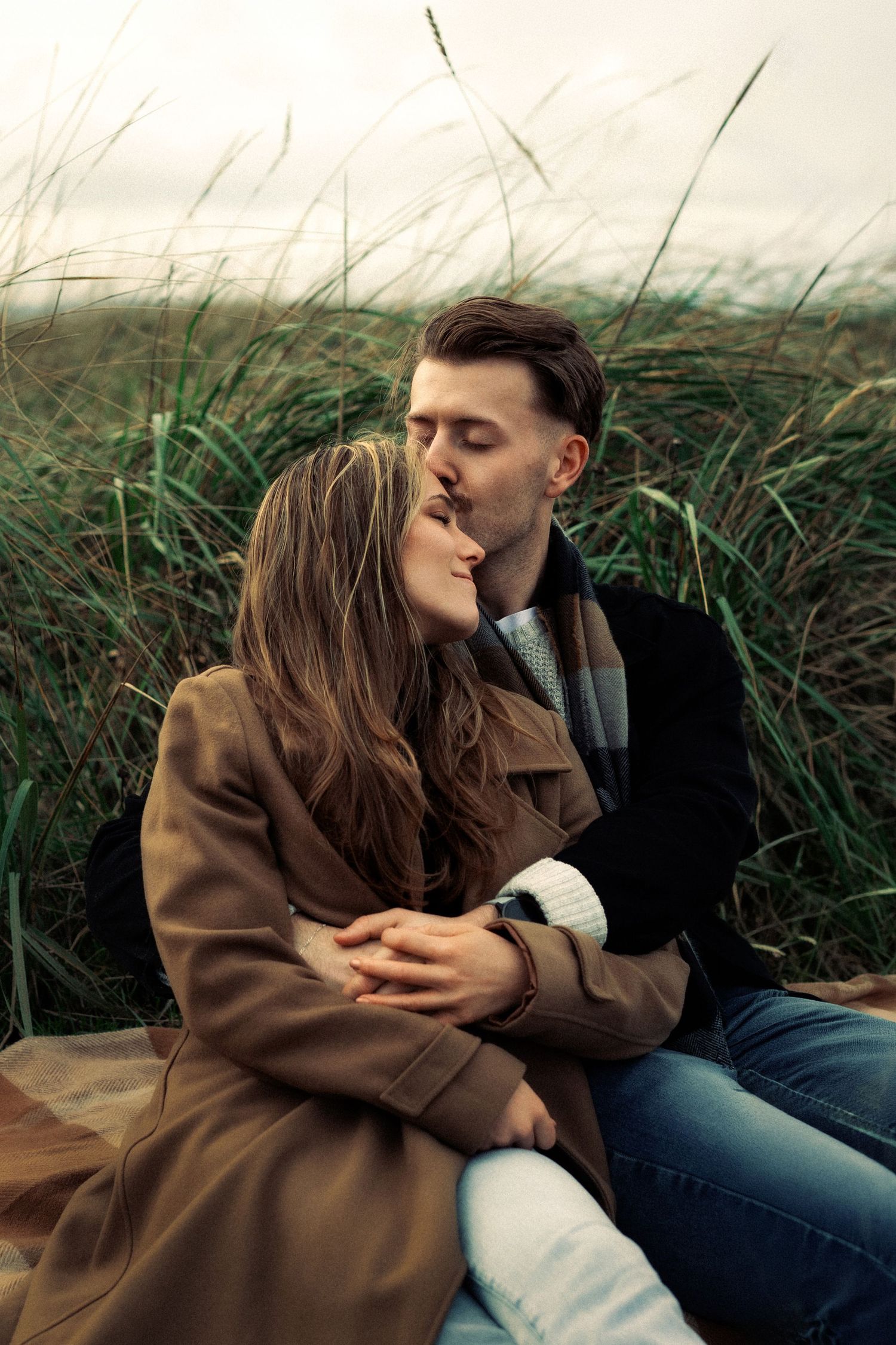 Romantic couple kissing outdoors in tall green grass, woman in brown coat embraced by man in dark jacket.