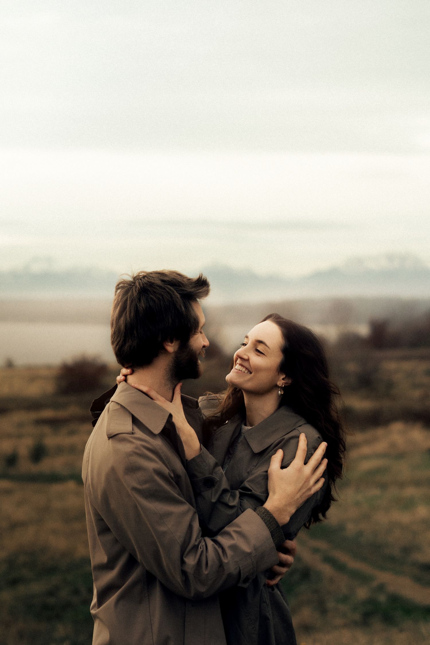 A couple embraces and smiles at each other in a misty outdoor landscape, both wearing matching olive trench coats.