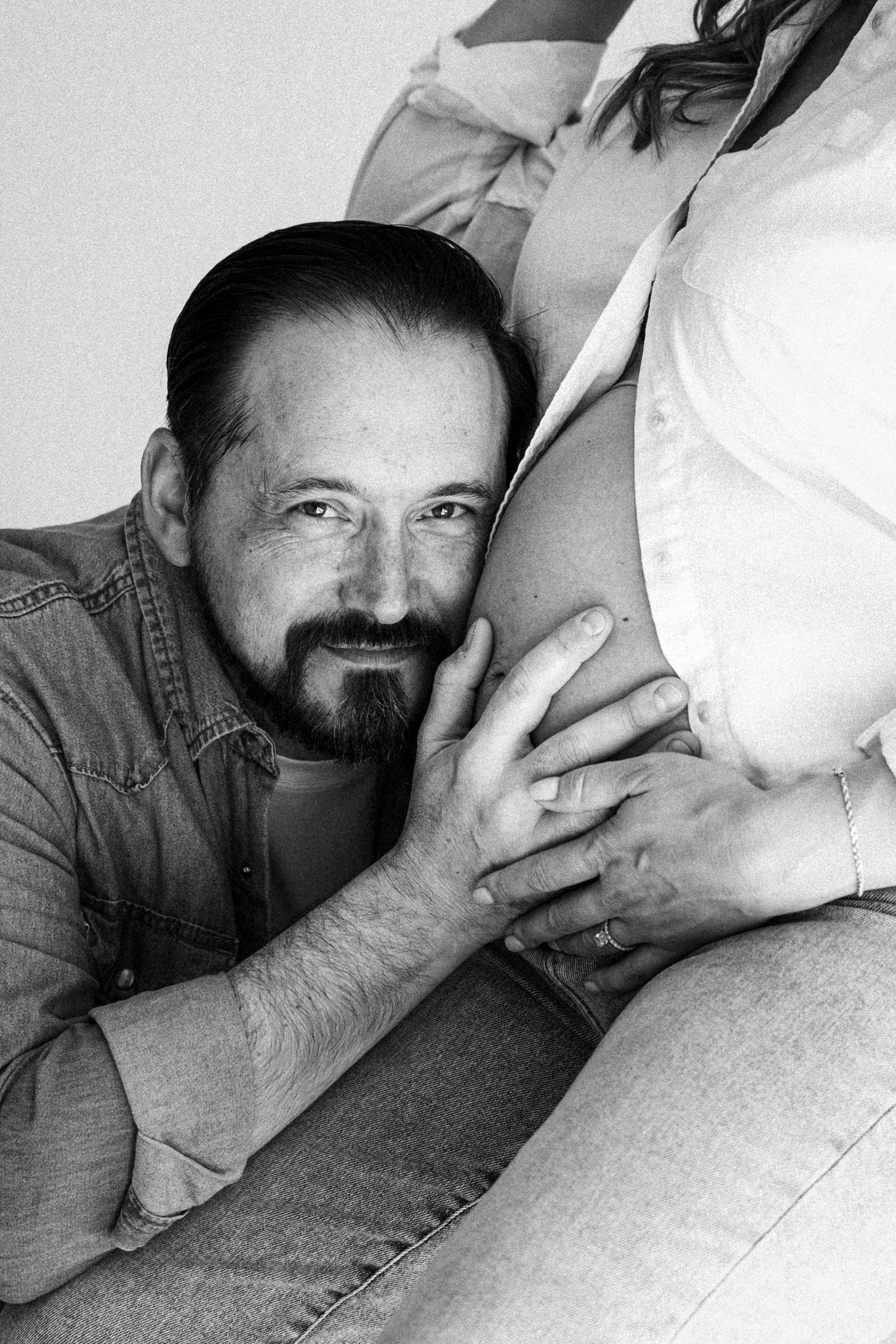 Black and white maternity photo of bearded man smiling while resting cheek against pregnant woman's bare belly.