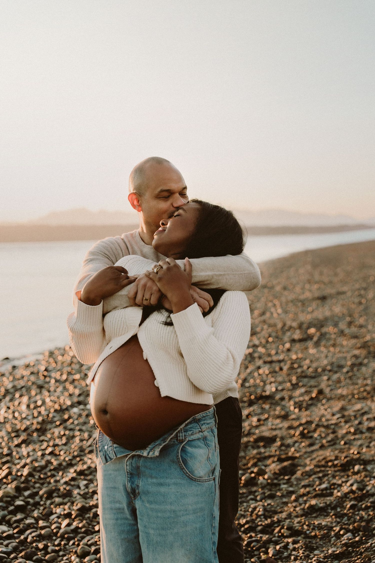 A couple shares a tender embrace on a rocky beach at sunset, the woman visibly pregnant, both smiling joyfully.